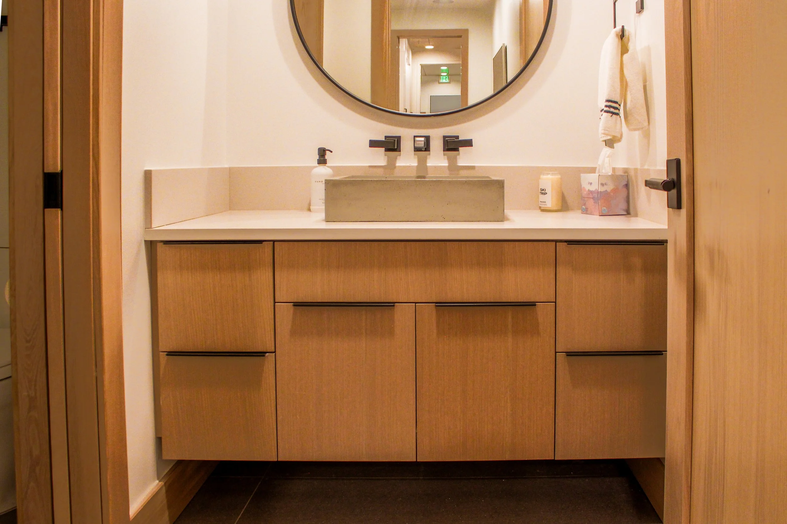 View of a bathroom vanity with a rectangular sink, a mirror above, two bottles of soap or lotion, a tissue box, and a towel on a hook. Wood cabinetry and beige countertops are visible.