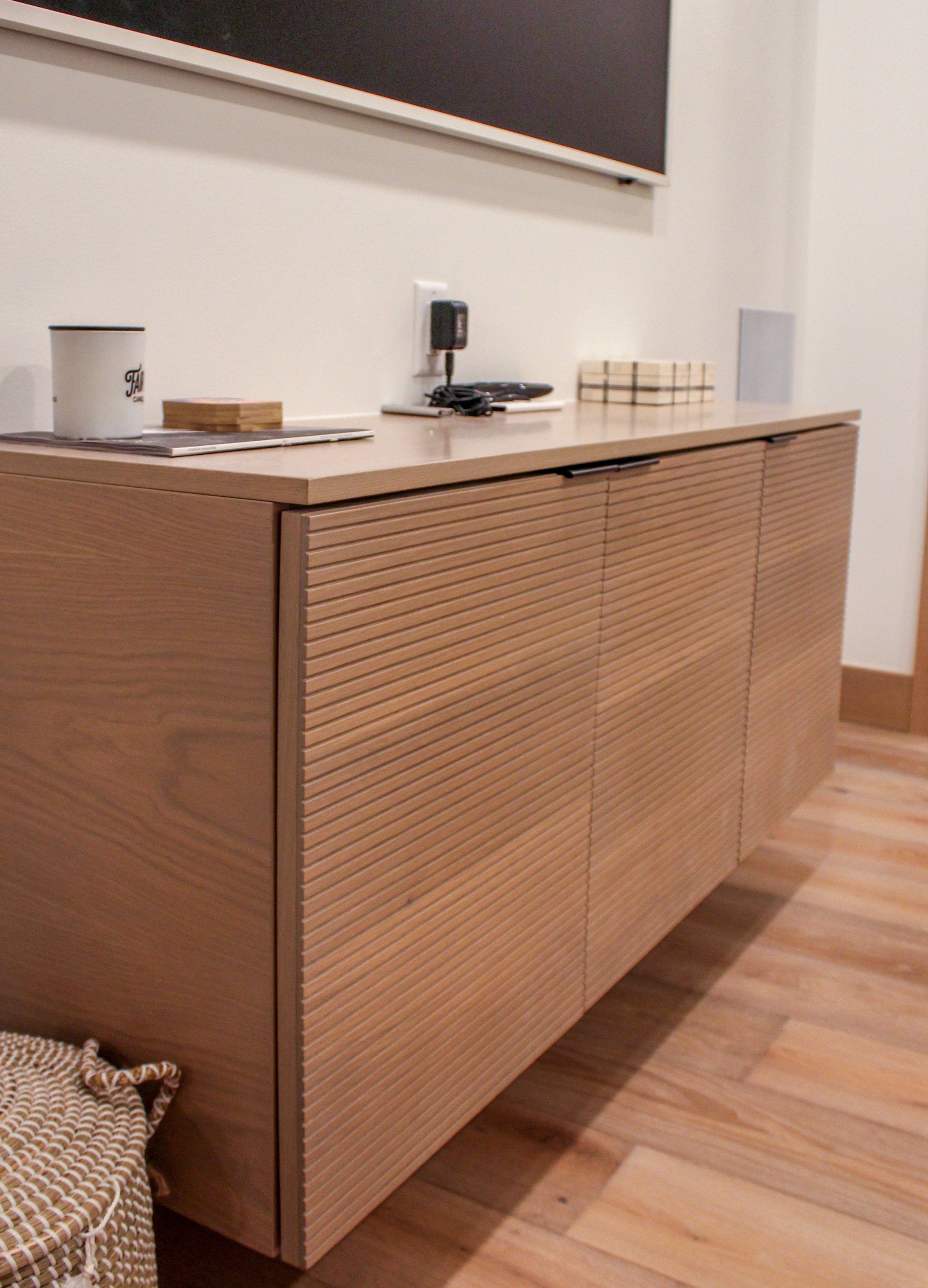 A wooden sideboard with closed doors in a room with wooden flooring. Items on top include a mug, a small wooden tray, a charging station with plugged-in devices, a stack of small boxes, and a small gray box. Part of a blackboard or chalkboard is visi