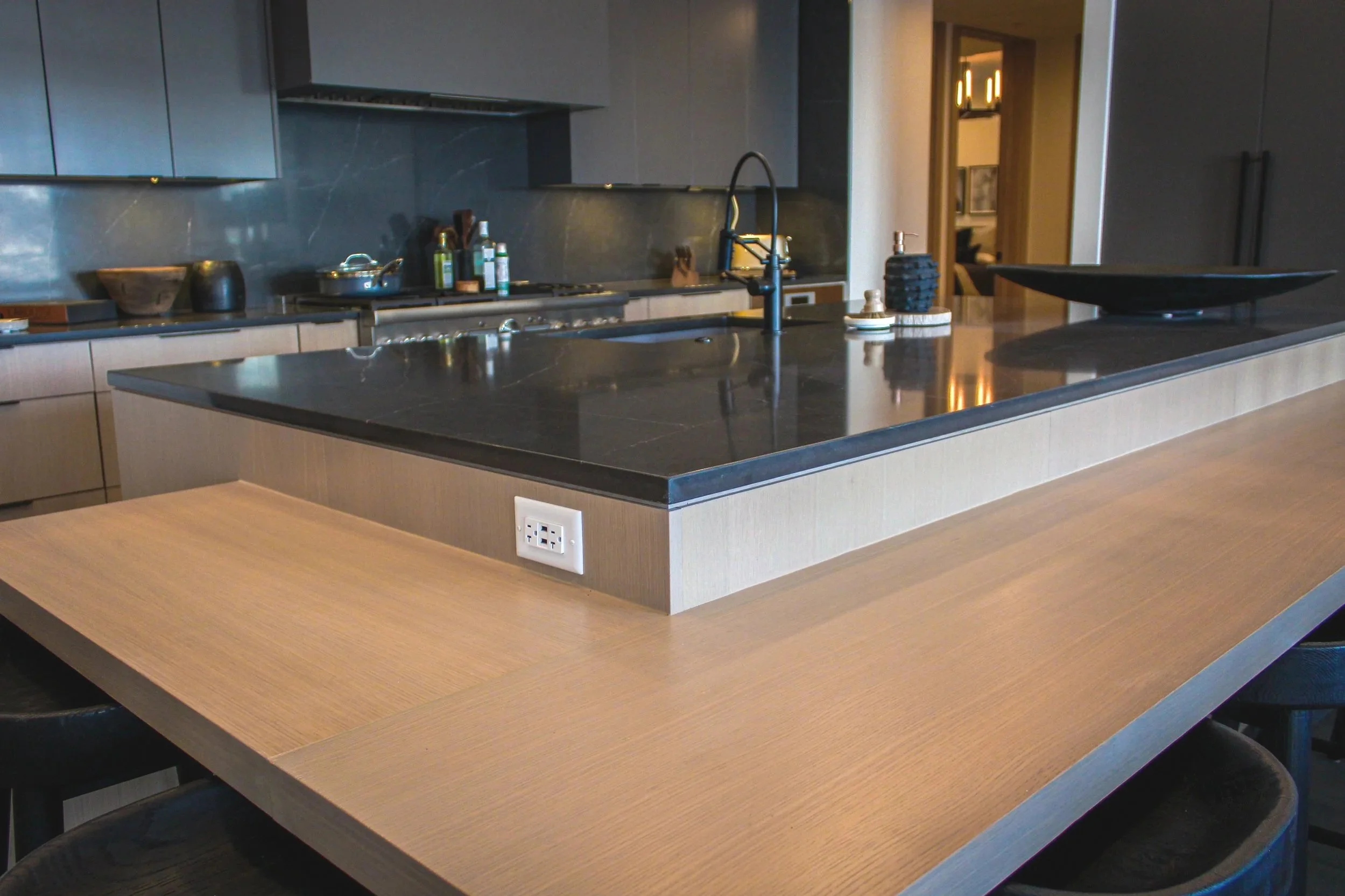 Modern kitchen with gray cabinets, black countertops, and a large island with a light wood finish. The kitchen faucet is black, and there are various bottles and kitchen items on the countertop. An electrical outlet is visible on the side of the isla