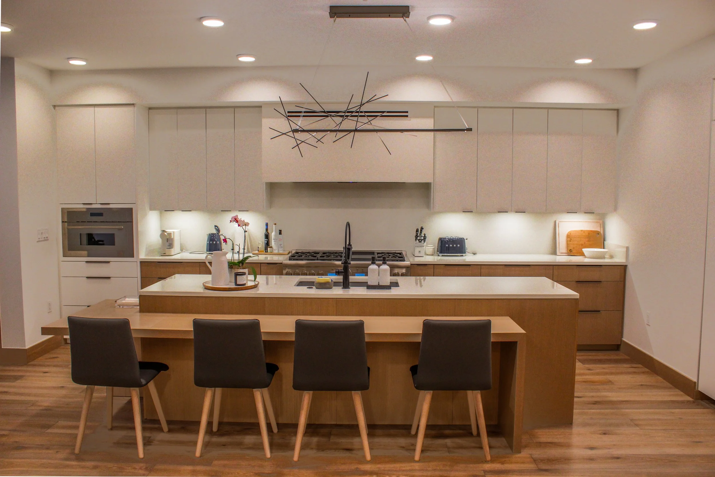 Modern kitchen with white cabinets, a center island, black chairs, and decorative black metal wall art, illuminated by recessed ceiling lights.