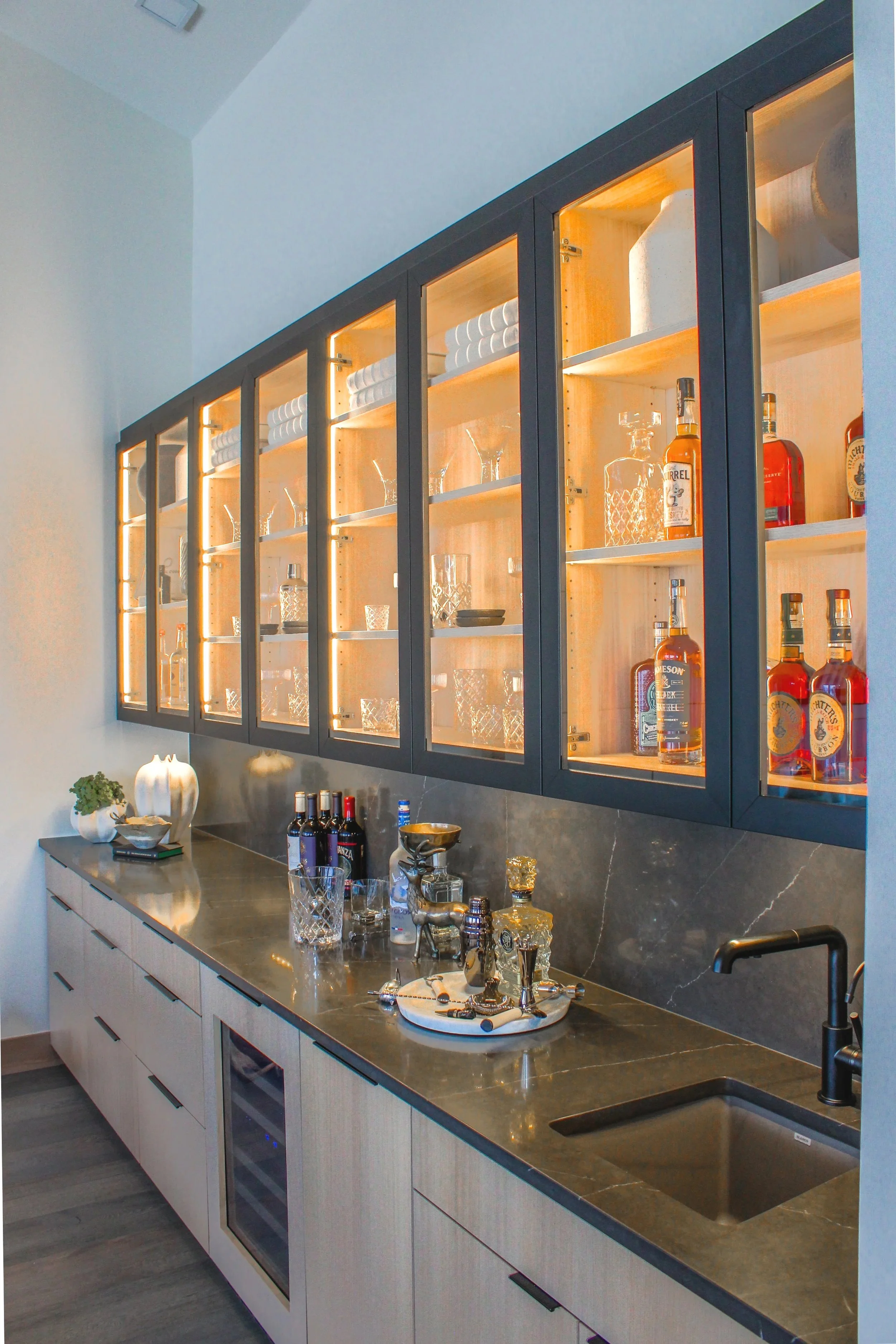 Modern kitchen with a black countertop, white cabinets, and a black glass-front wine cooler. Open shelving behind glass doors displays liquor bottles, glassware, and decorative items. On the counter, there are bottles of wine, a tray with jewelry, a 