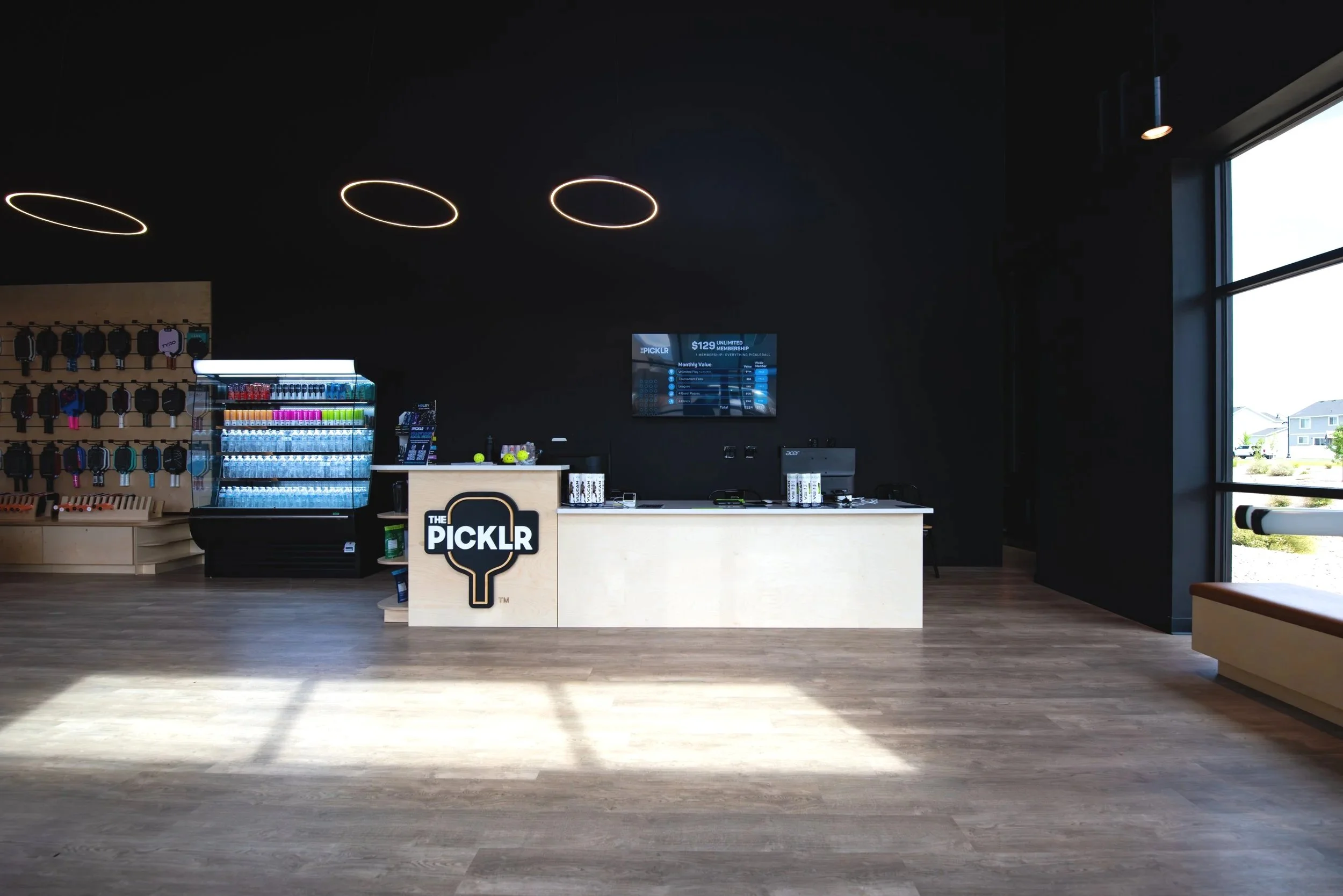 Interior of a modern sports retail store with a black wall, illuminated ring ceiling lights, a refrigerated display of drinks, shelves with sports accessories, and a checkout counter with a digital screen.