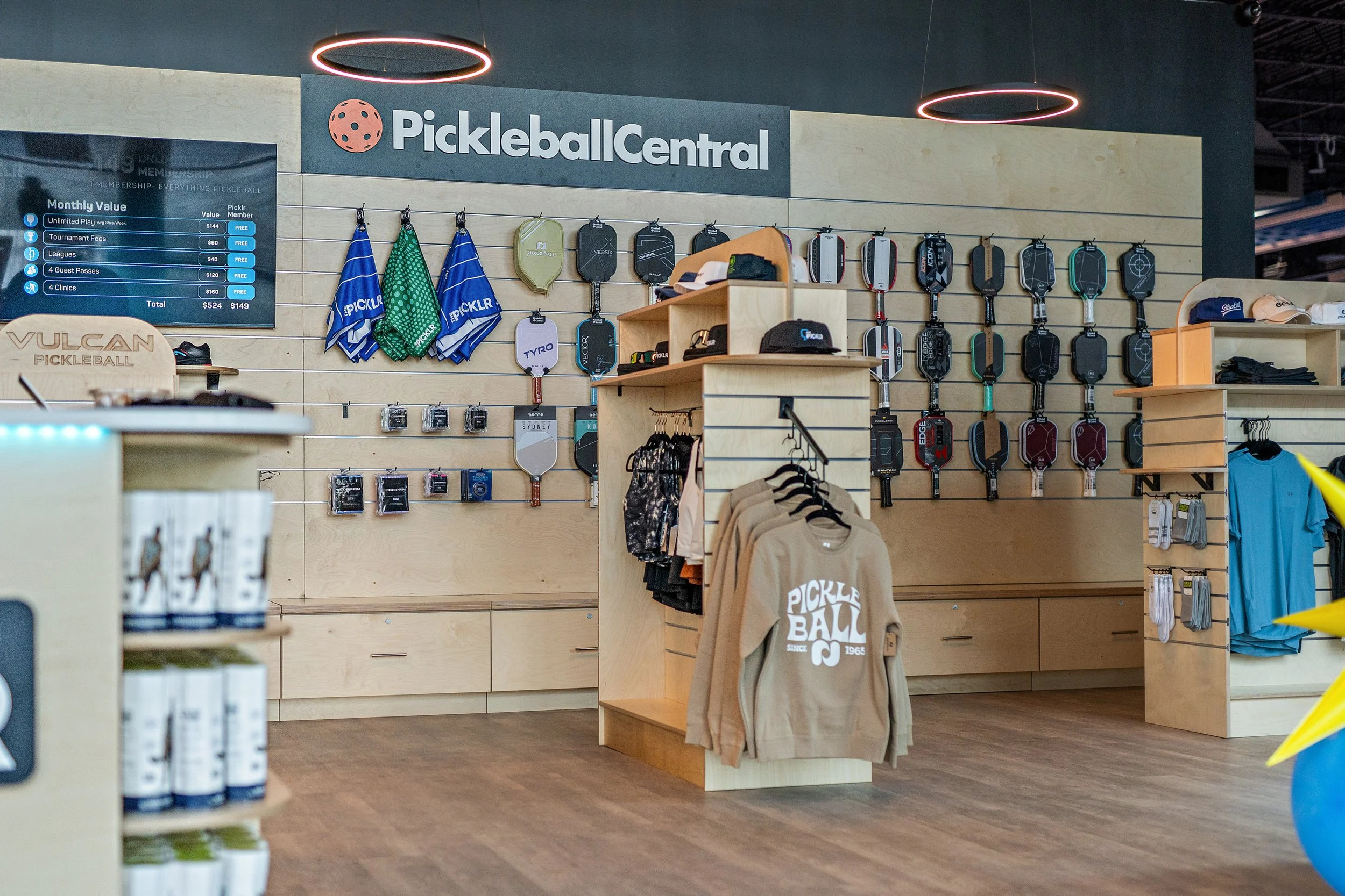 Interior of a retail store called Pickleball Central, displaying pickleball paddles hanging on the wall, merchandise shelves, and clothing. The store has modern lighting and wooden display fixtures.