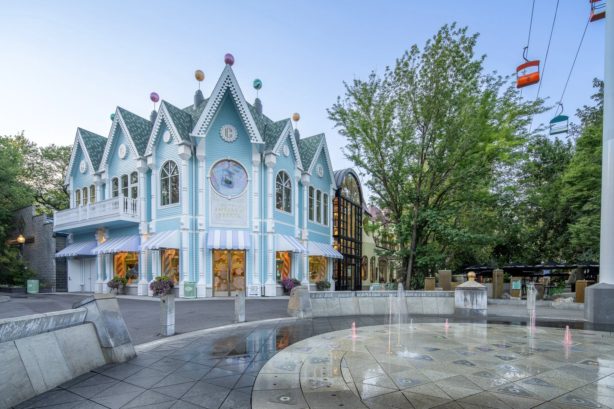 A blue and white Victorian-style building with a sign that reads 'Sweet Treats,' located in an amusement park or fairground. There is a fountain with pink jets of water in the foreground and a large tree to the right. A cable car or gondola lift with colorful seats is visible on the right.