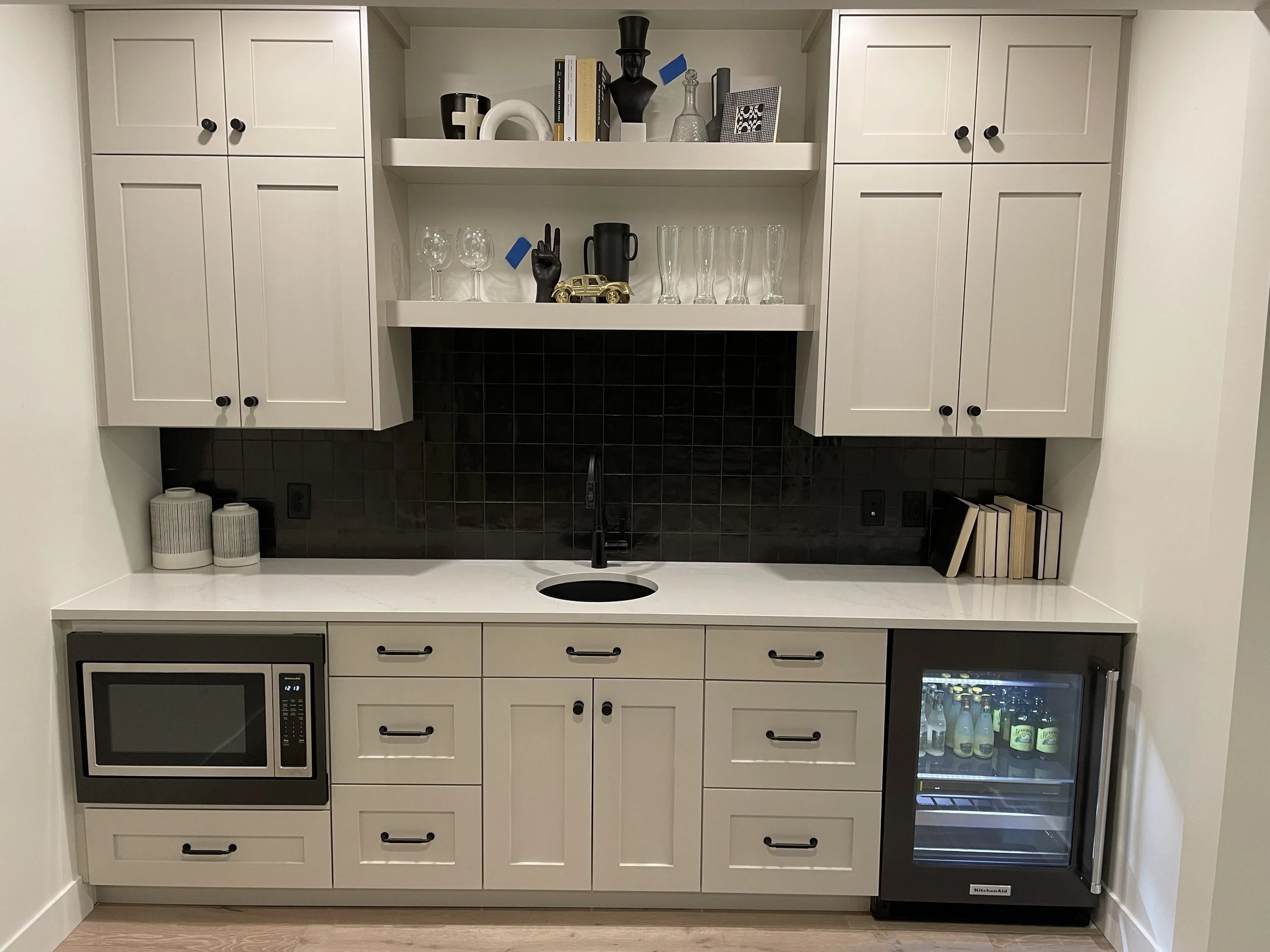 A modern kitchenette with white cabinets, black hardware, a black tiled backsplash, and a white countertop. There is a microwave and a mini fridge built into the lower cabinets, a wine cooler on the right side, and decorative items and glassware on the open shelves above. The area is well-lit and clean.
