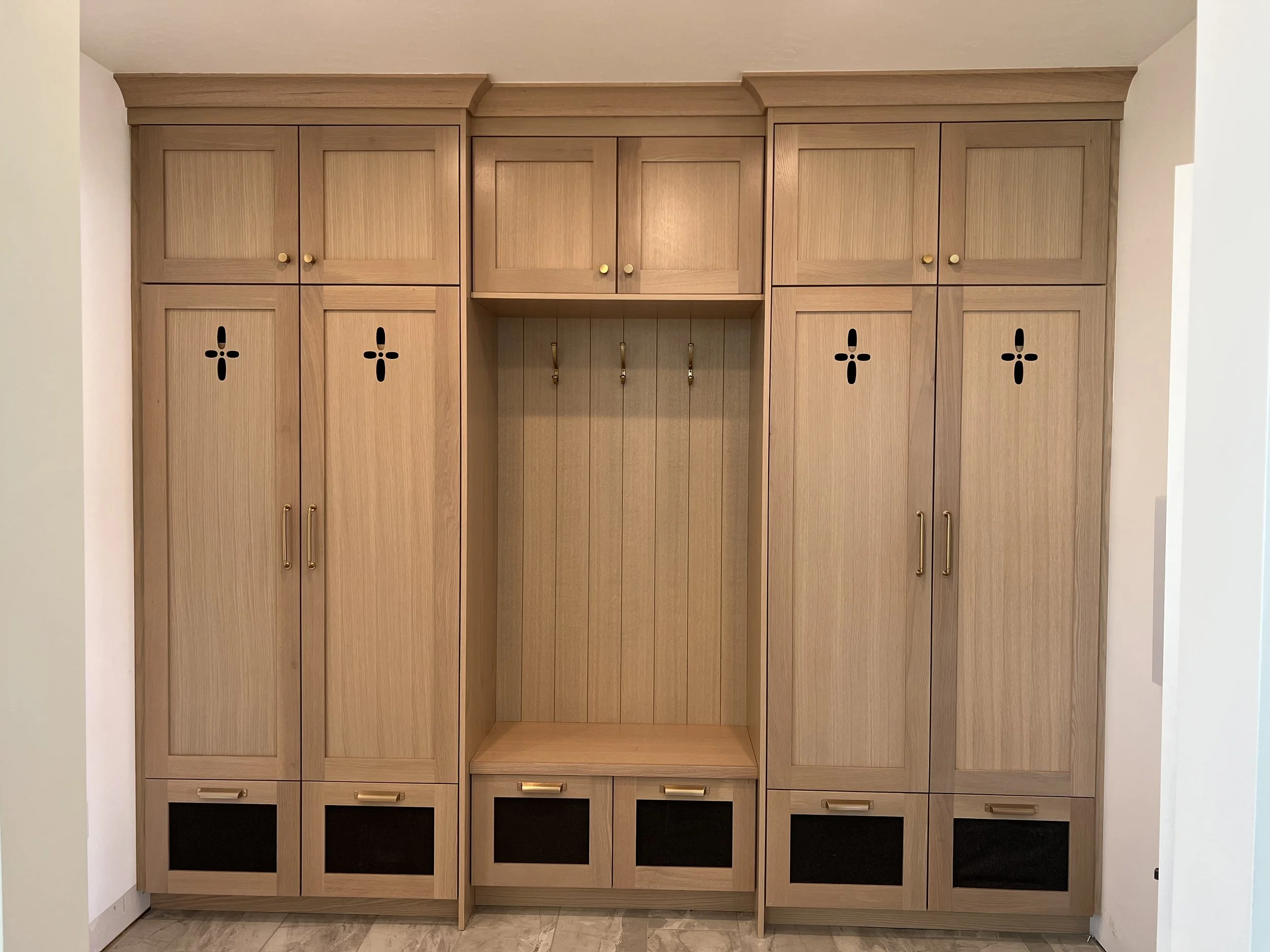 Wooden cabinet with multiple doors and golden knobs, including a central open section with hooks, and cubbies at the bottom, set against white walls.