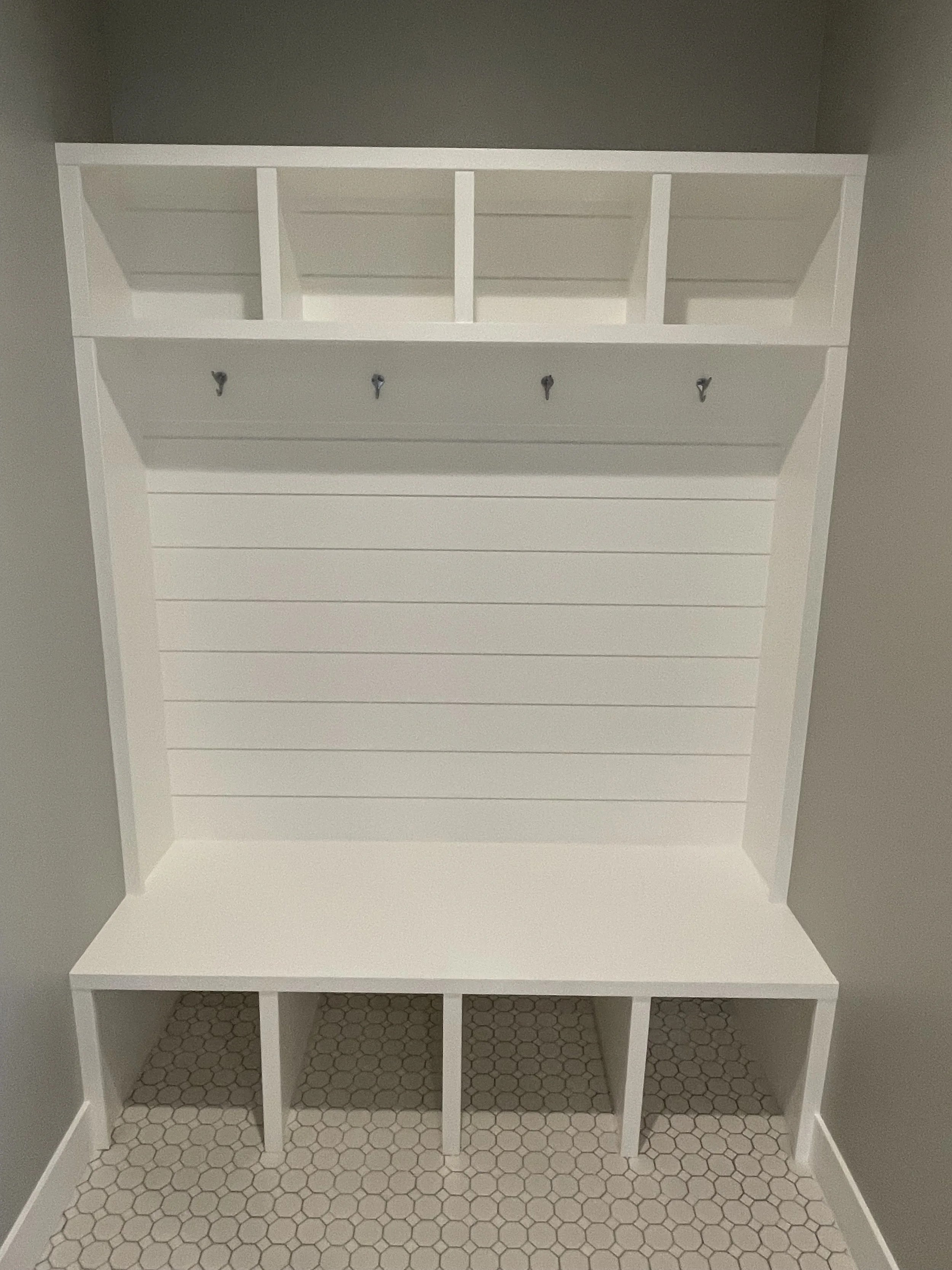 Empty white wooden cubby shelf with hooks and divided compartments, situated against a gray wall with hexagonal tile flooring.