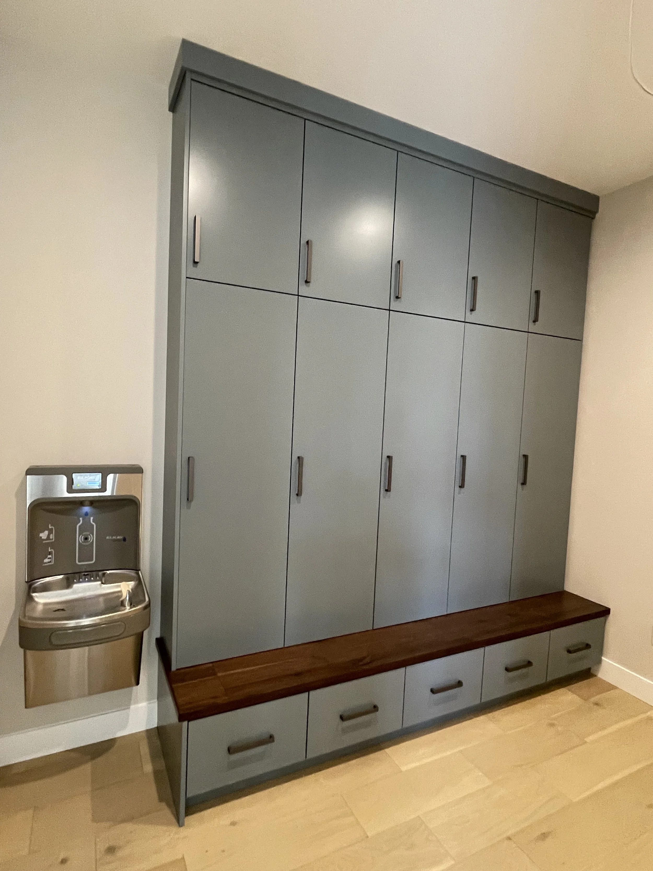 A blue wall-mounted cabinet with multiple compartments and a wooden bench attached at the bottom, next to a water fountain on the wall.