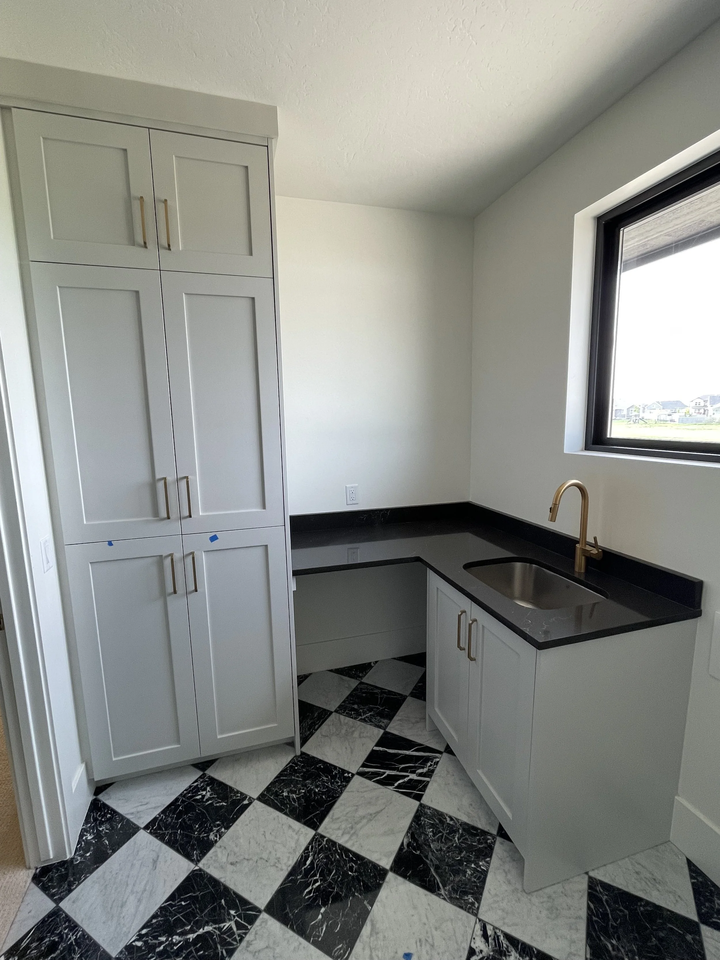A modern kitchen with black and white checkered marble floor, white cabinets with gold handles, black granite countertop, and a black sink under a window showing a view of a neighborhood.