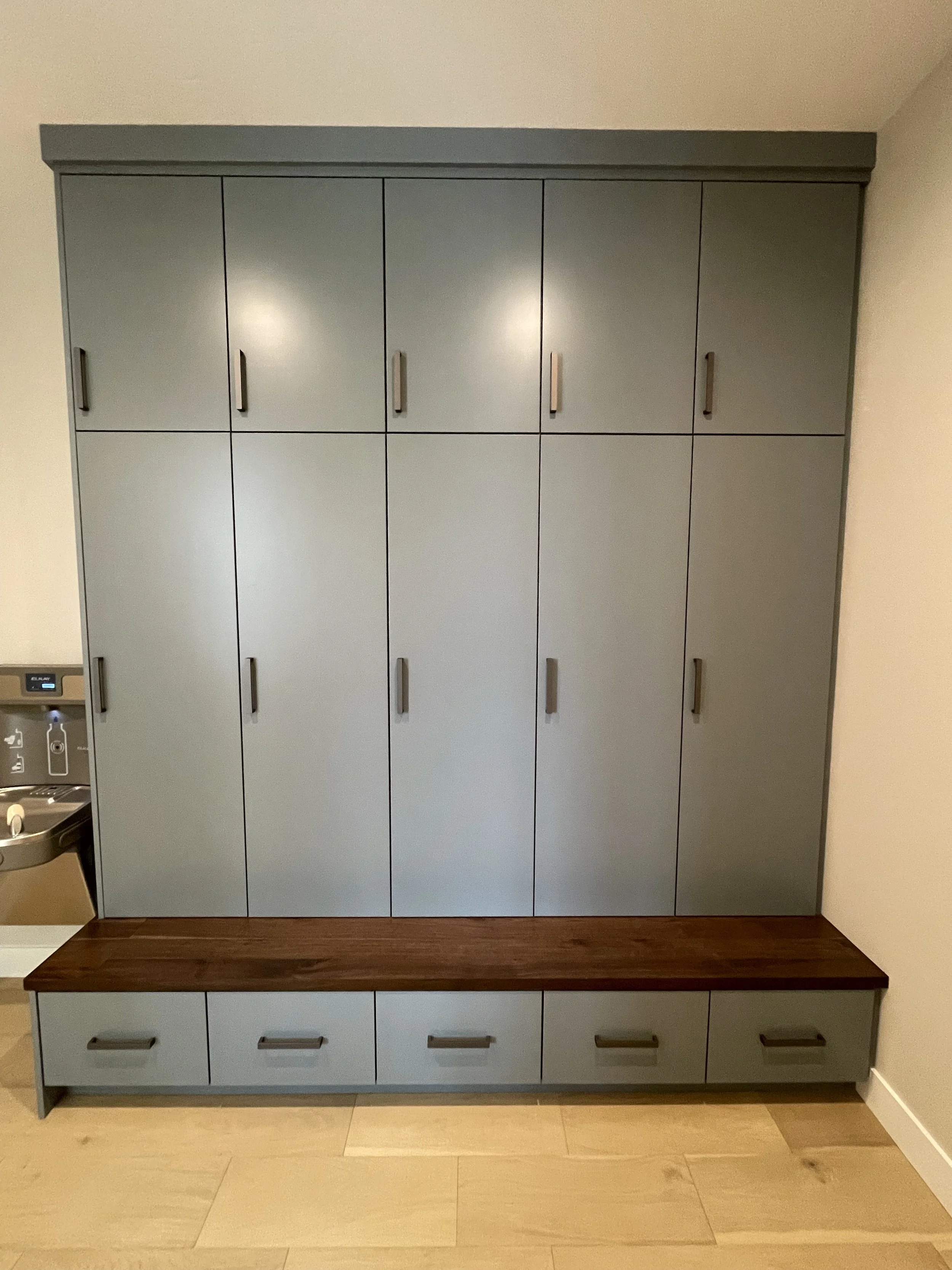 A blue and gray storage cabinet with multiple doors and drawers, situated against a white wall on a wooden floor, with a small bench in front and a water fountain to the left.