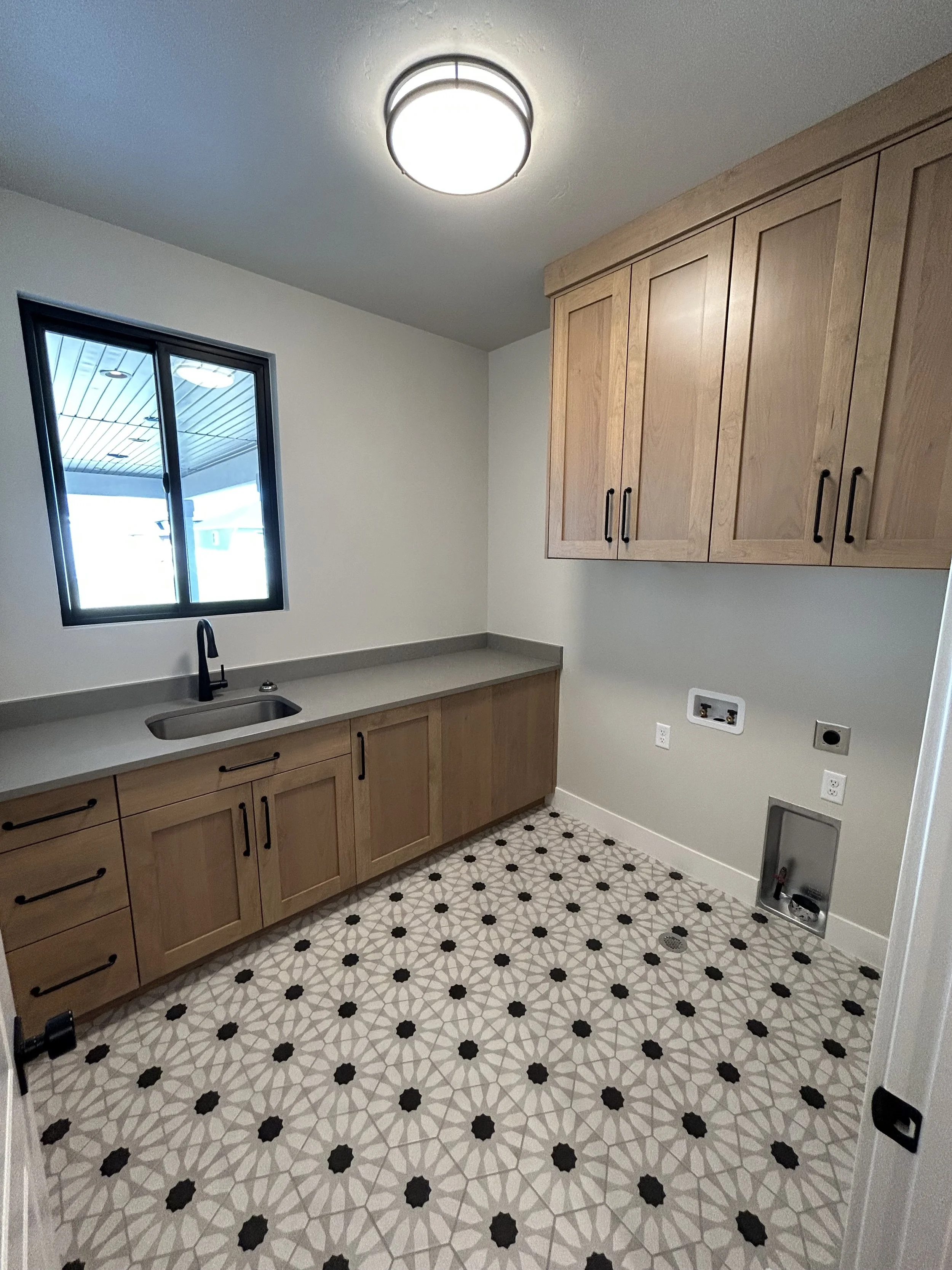 Empty kitchen with beige cabinets, a gray countertop, a small black faucet and sink, tiled patterned floor, and a window on the wall showing an outdoor covered area.