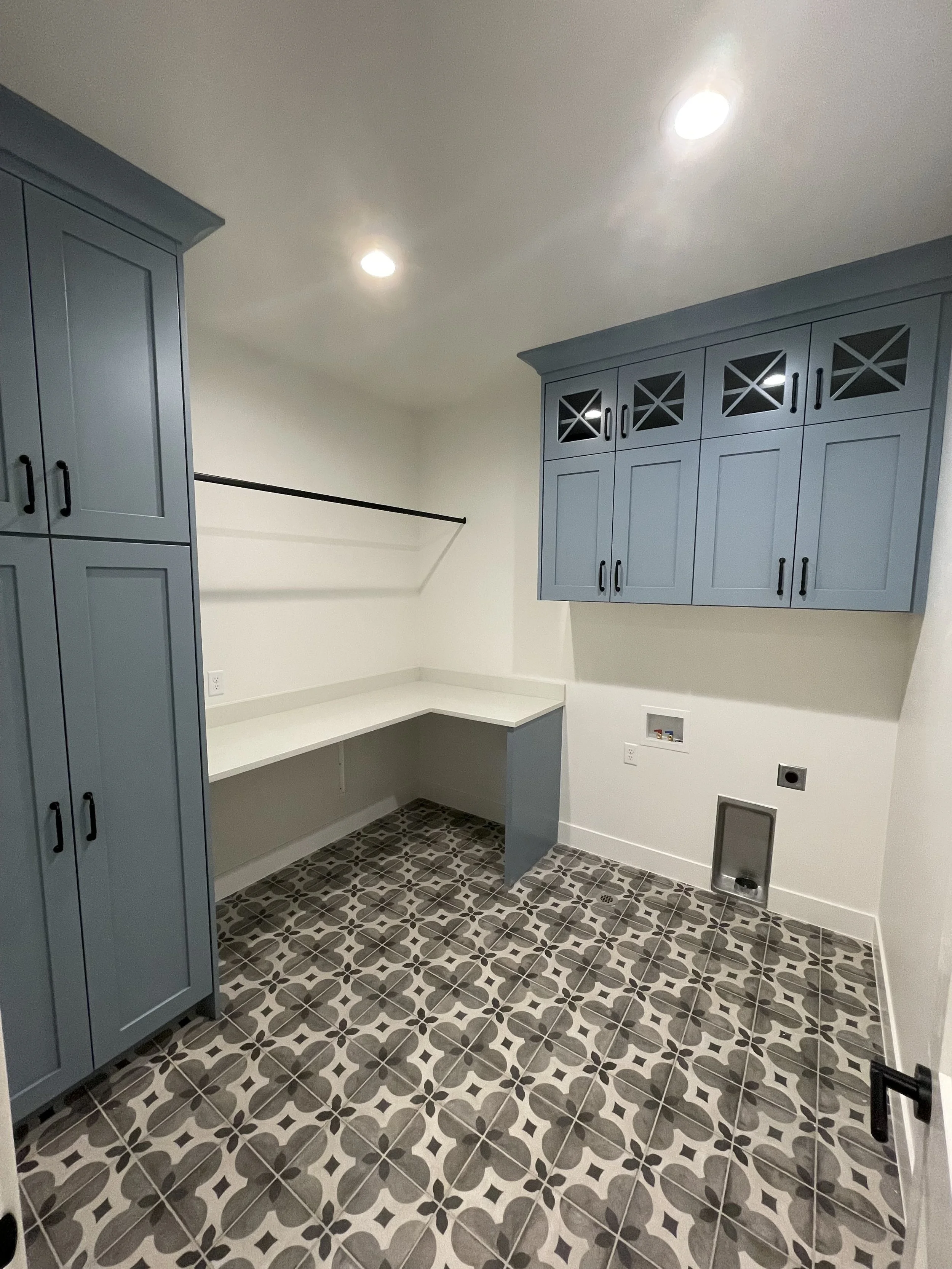 An empty laundry room with dark patterned tile floor, white walls, blue cabinets, and a white countertop with open space underneath.