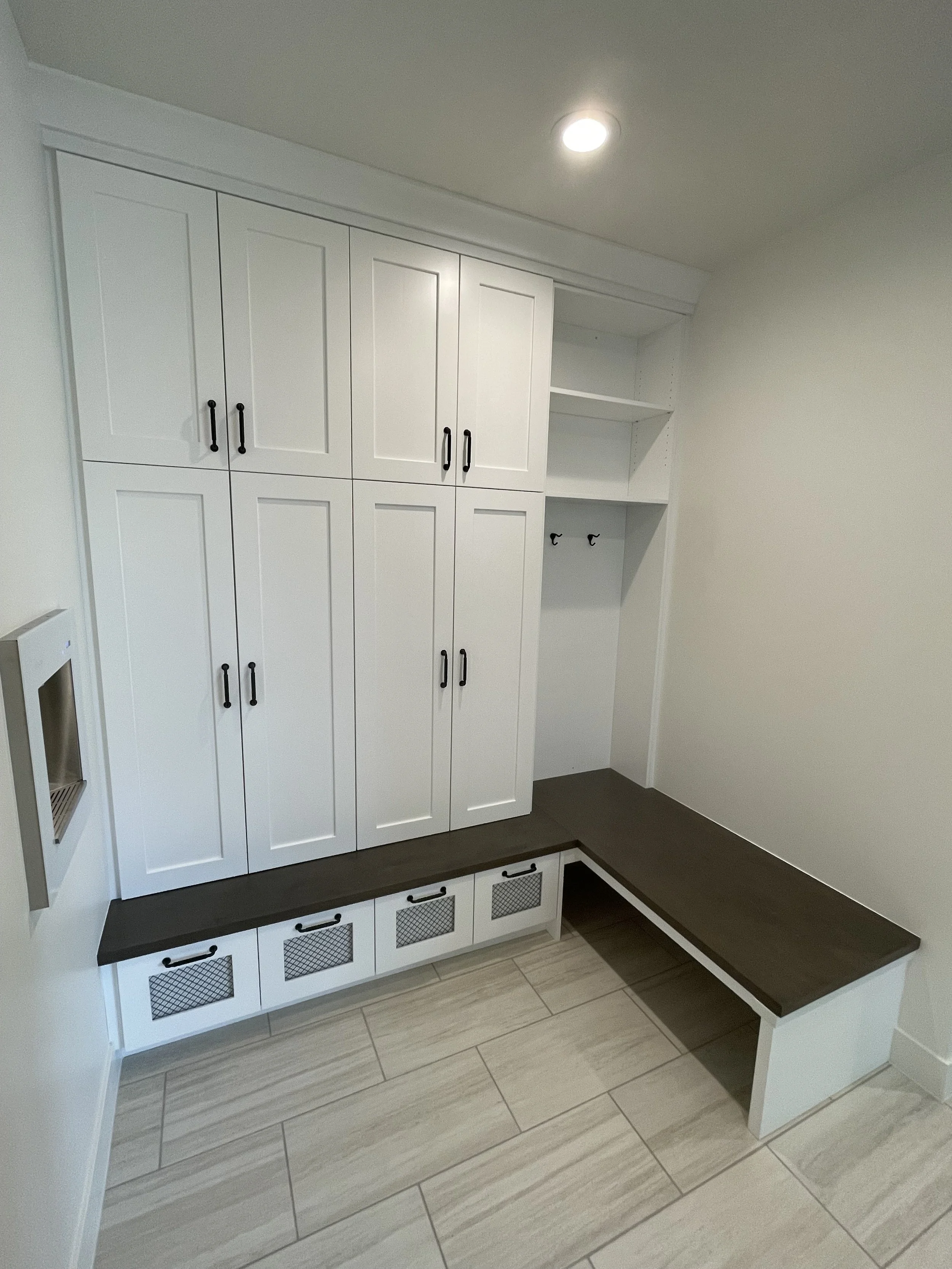 A white kitchen cabinet with black handles, a black countertop, and multiple Storage compartments with mesh fronts. There is a small countertop space with a bench attached to it, and the floor is tiled with light-colored tiles.