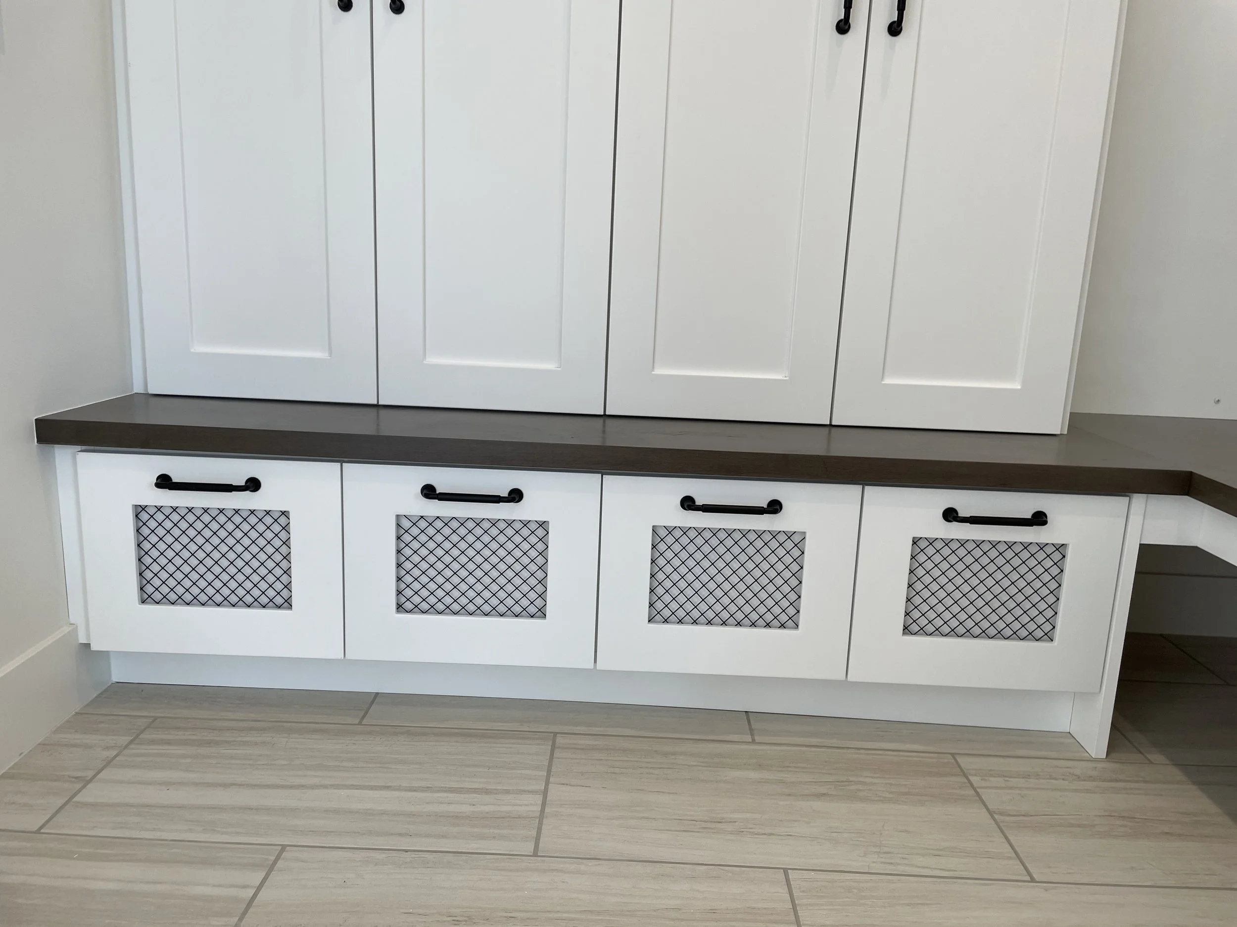 A white cabinet with a dark brown countertop, black handles, and decorative grille panels, set against a wall with beige tiles and a wooden floor.