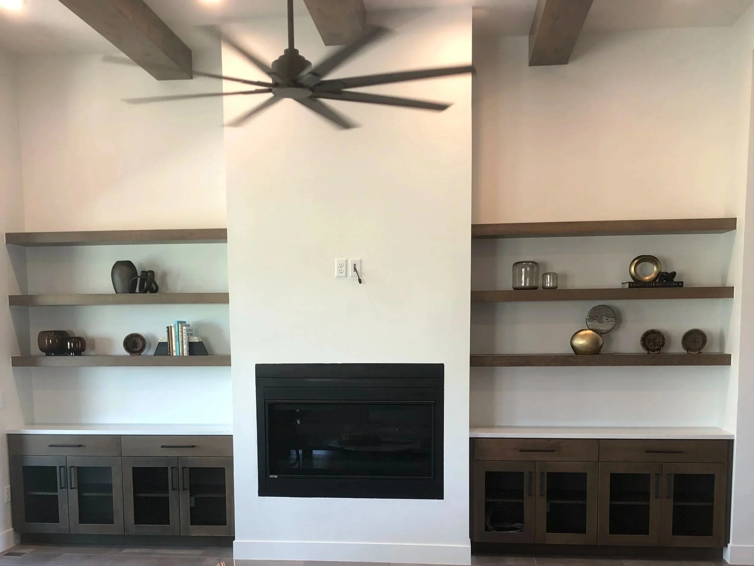 Interior living room with beige walls, wooden beams, floating shelves with decorative items, a black fireplace, and a ceiling fan.