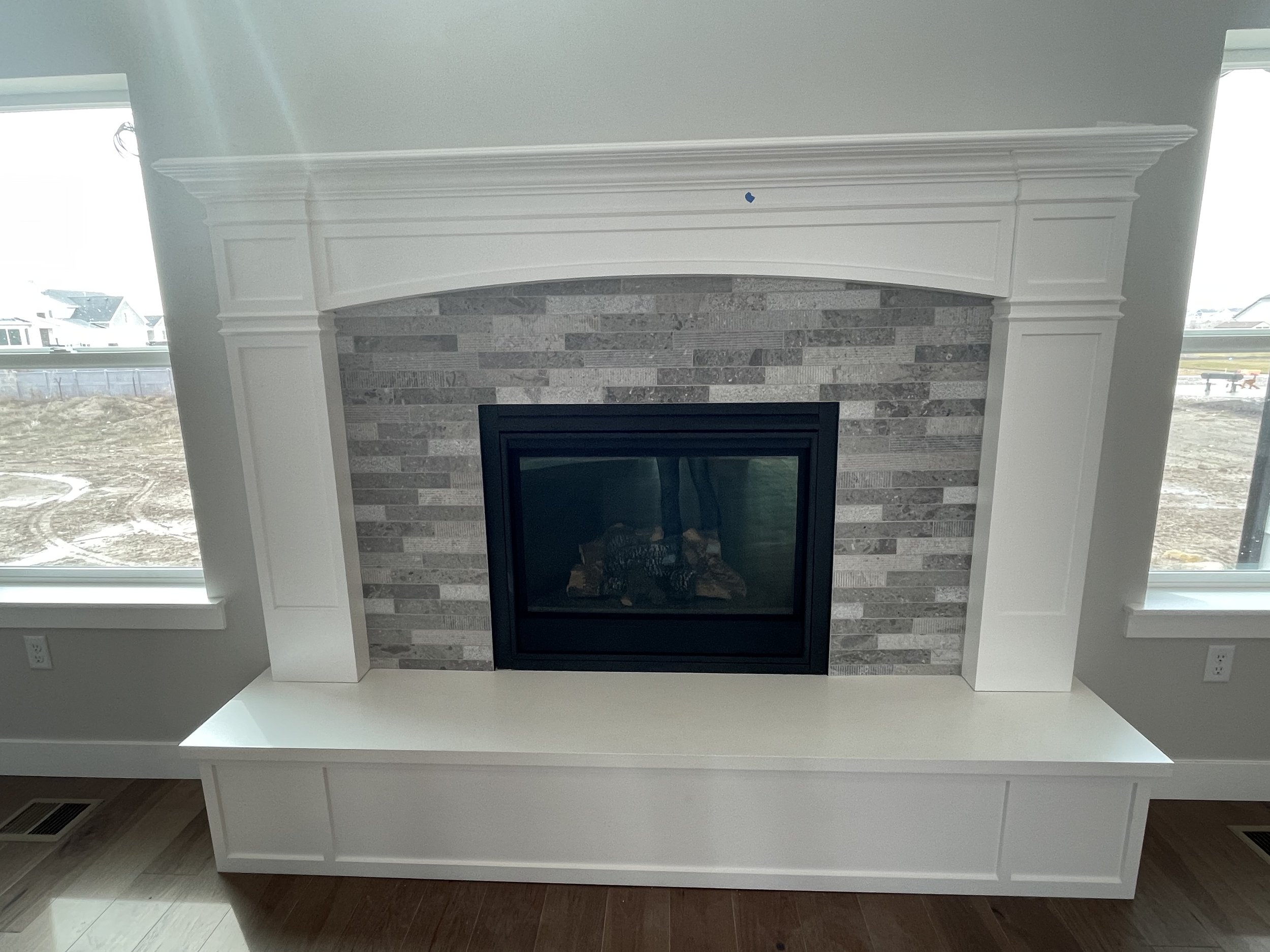 White fireplace mantel with a stone surround, situated between two large windows in a living room, during daytime.