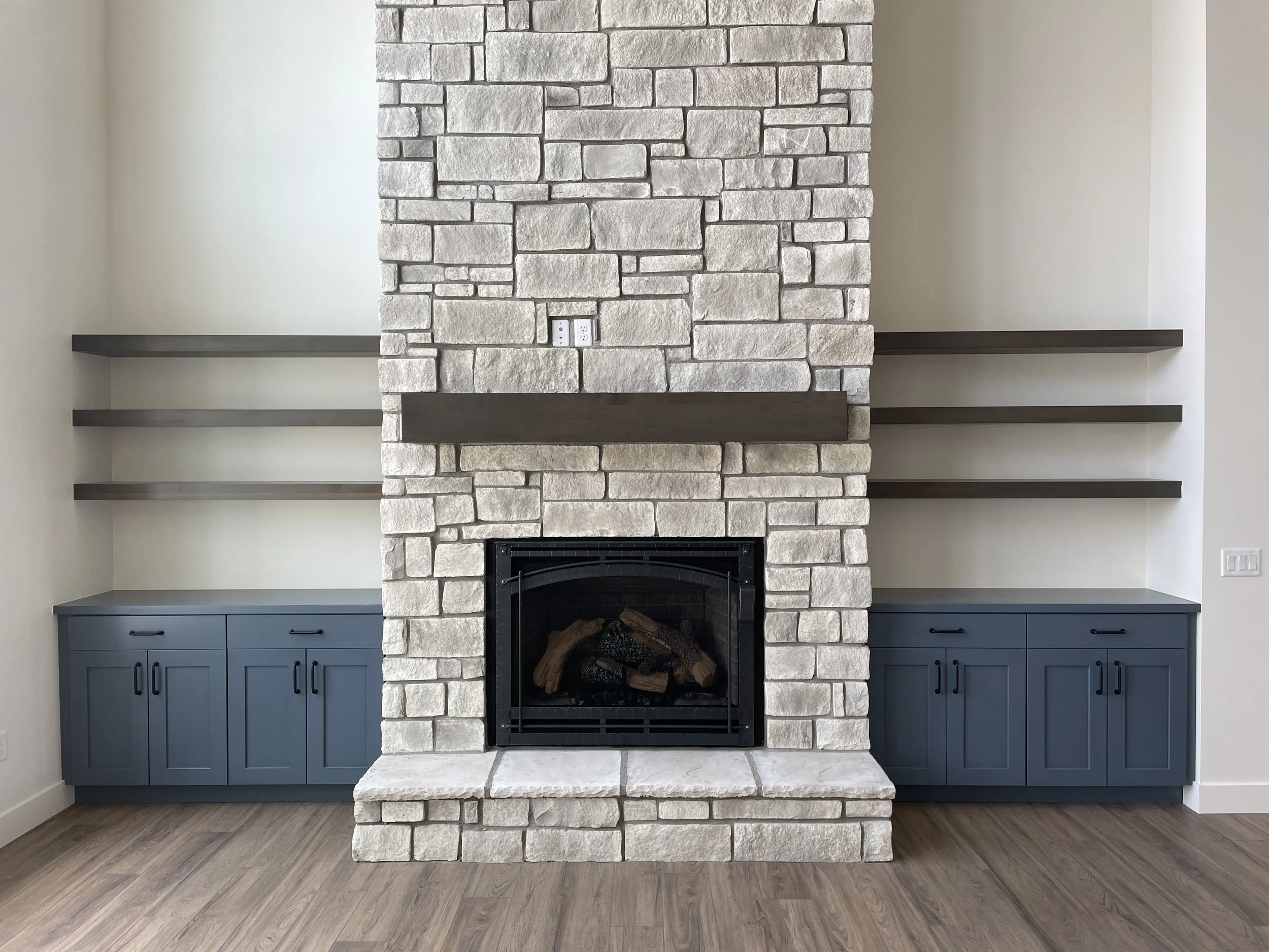 A modern fireplace with a stone surround, flanked by blue cabinets with black handles, and two floating shelves on either side. The fireplace has a metal frame with logs inside, and the room has light-colored walls and wooden flooring.