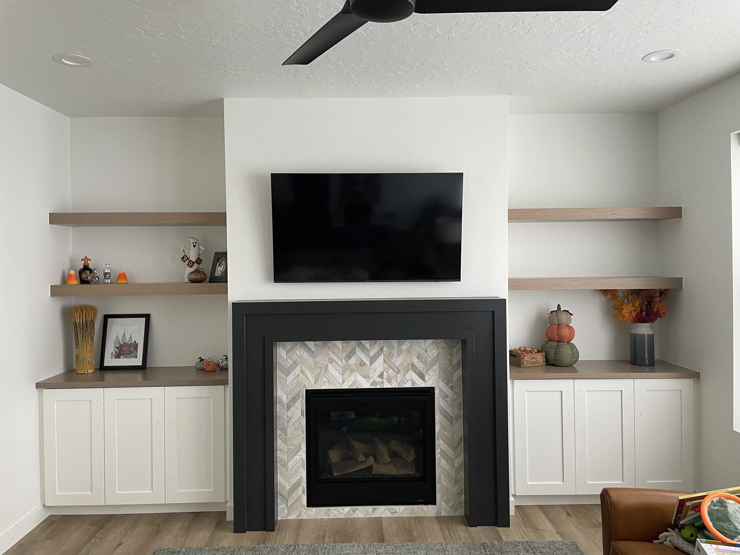 Living room with a fireplace, white cabinets, wooden shelves decorated with small pumpkins, a framed picture, and a TV mounted above the fireplace, with a white wall and wood-look flooring.