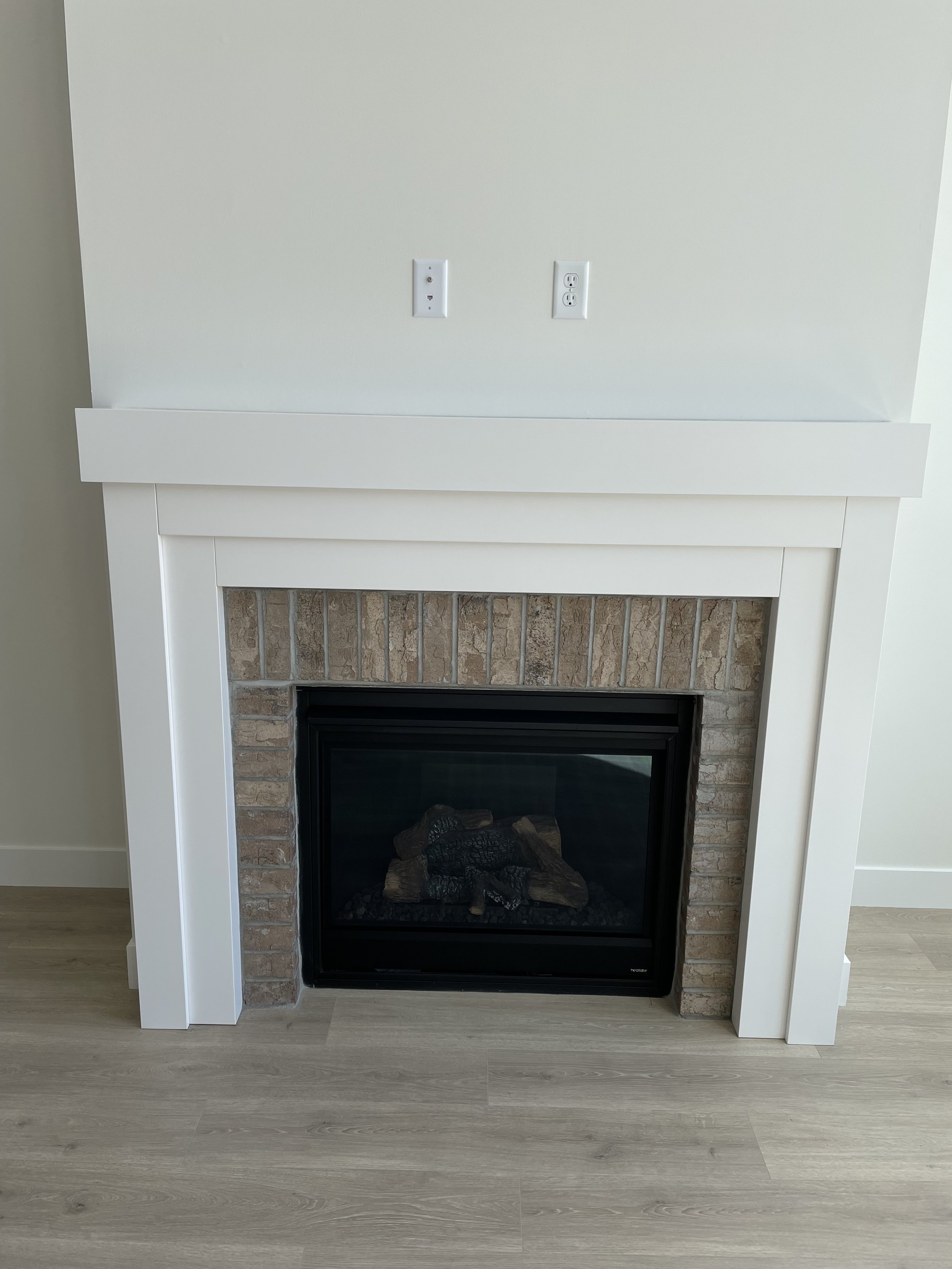 Empty living room fireplace with a white mantel, brick surround, and a black framed glass front, along with outlets and a wall pipe above.