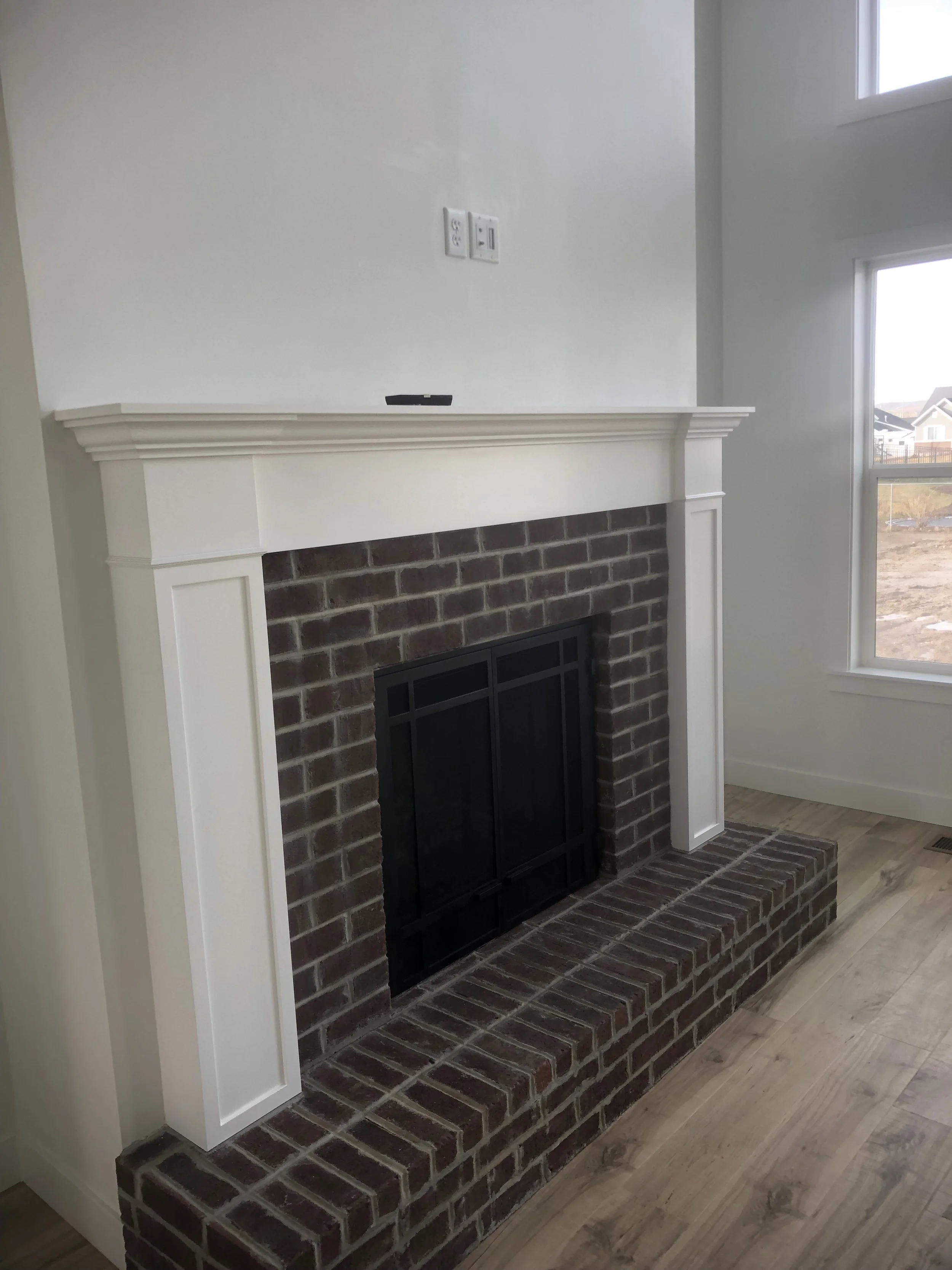 Living room with brick fireplace, white mantel, two electrical outlets, and a large window showing a suburban neighborhood outside.