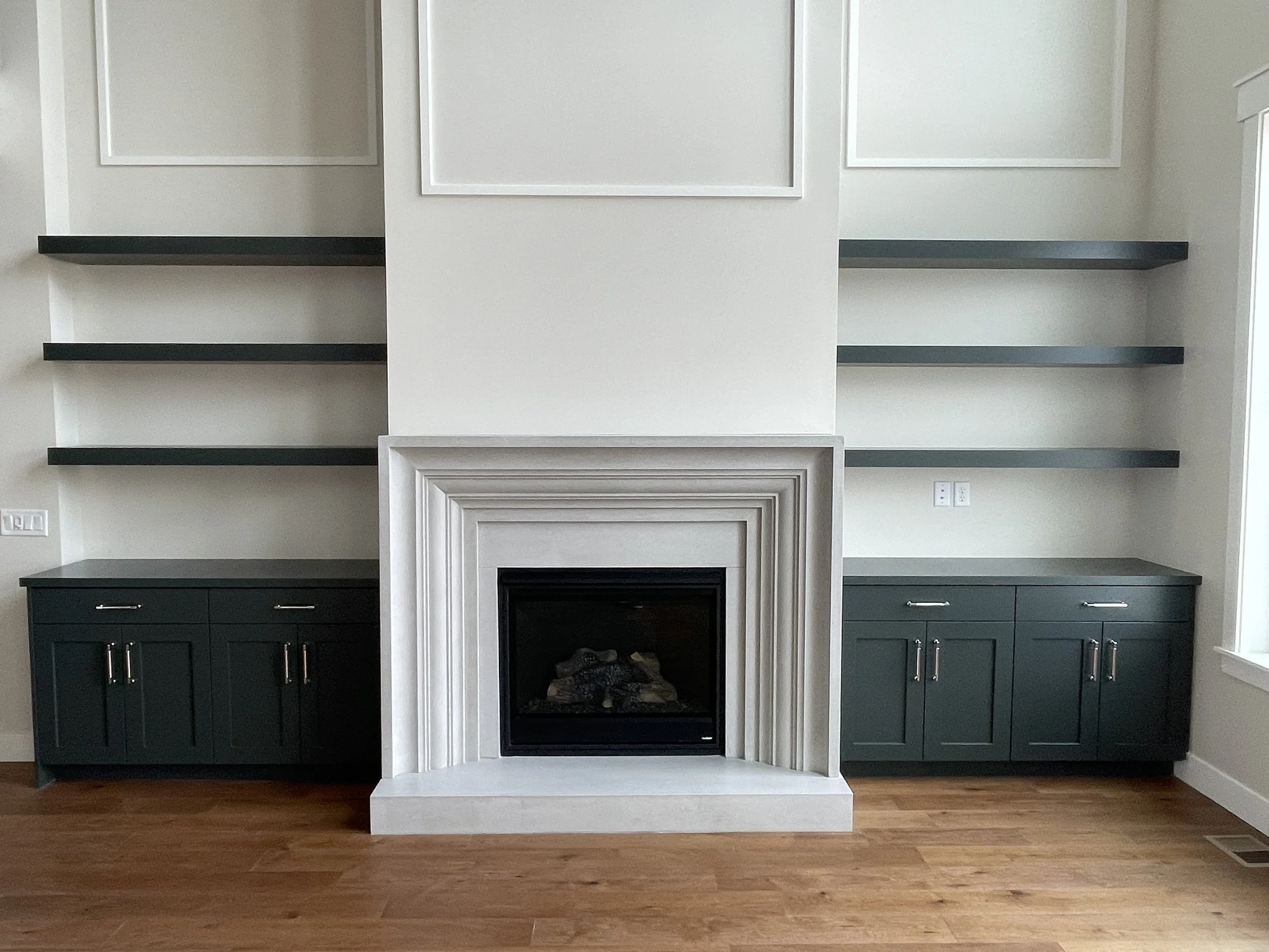 Empty built-in fireplace with white trim, flanked by black cabinets with silver handles, and open shelves above, in a white wall room with hardwood flooring.
