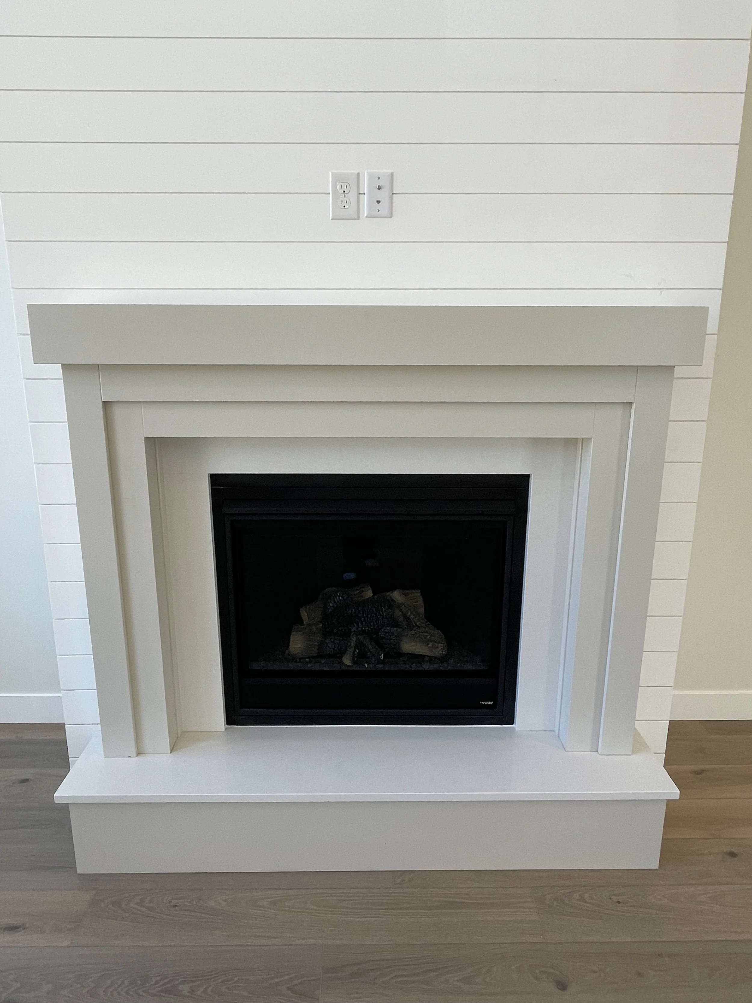 White fireplace with a black interior, situated on a wood floor, with a white mantle and wall paneling above.