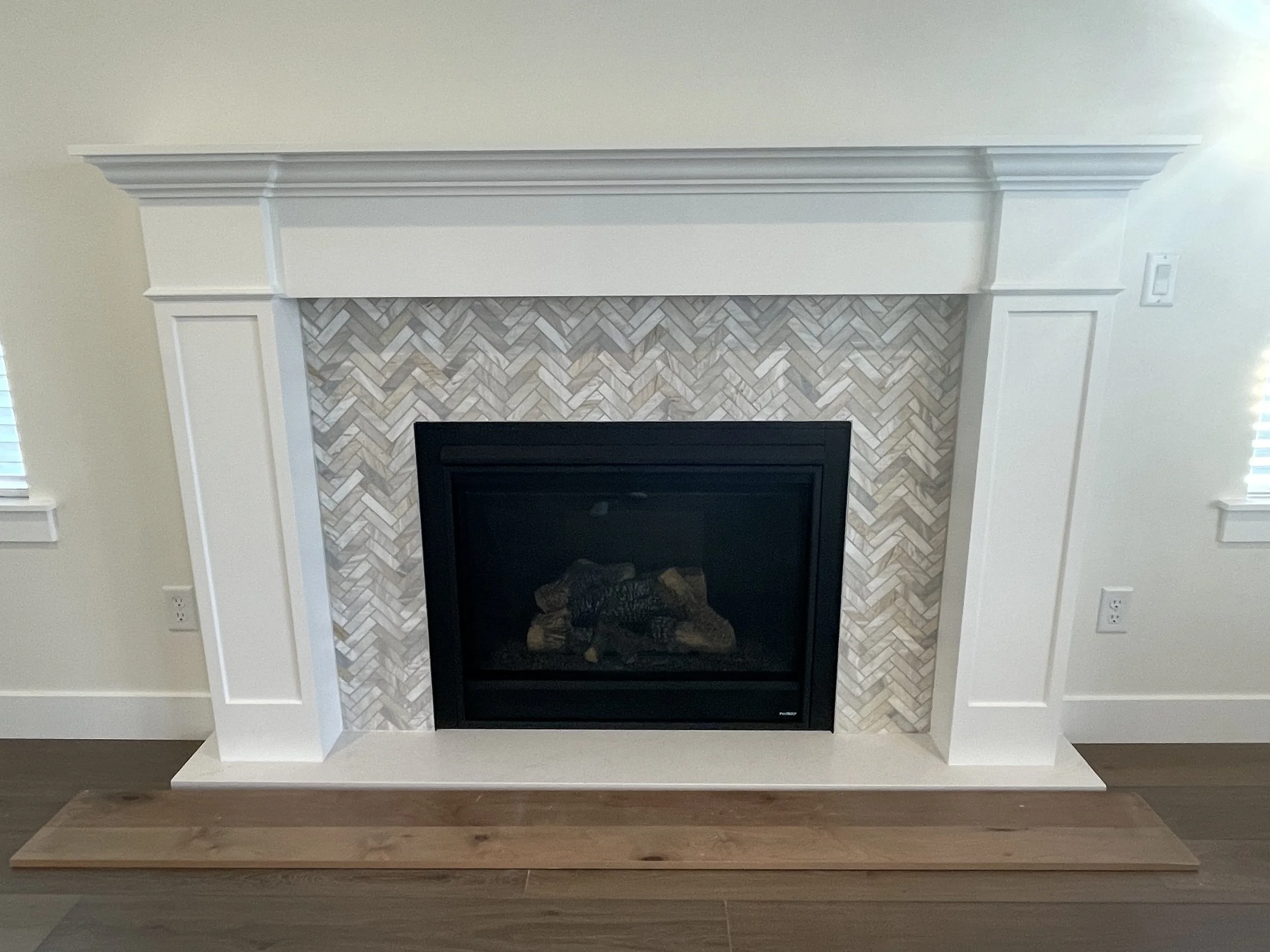 A fireplace with a herringbone tile surround, black trim, white mantel, and decorative molding on either side.