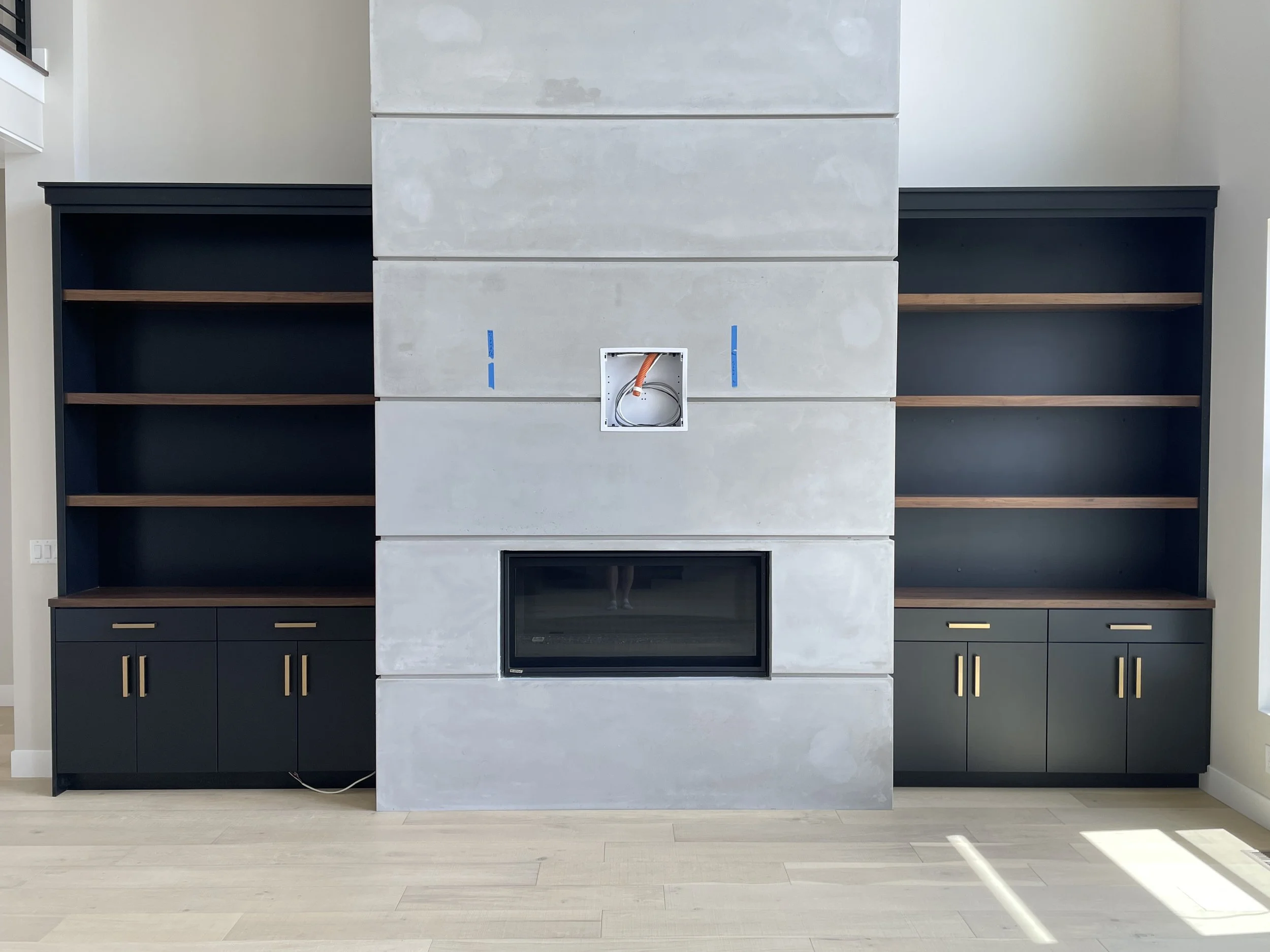 Empty built-in entertainment center with a fireplace at the bottom, surrounded by open shelving on both sides, and a central section with an electrical box and wiring, in a modern living room.