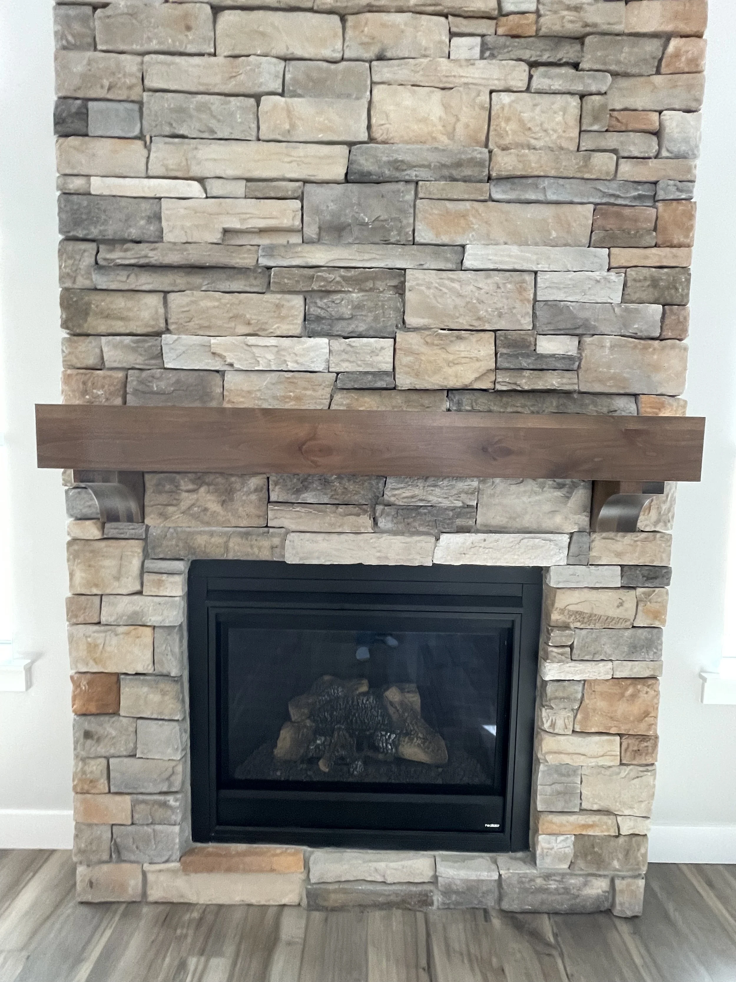 A fireplace with a black framed glass opening, surrounded by a beige, gray, and tan stacked stone facade. A wooden mantle extends across the top of the fireplace.