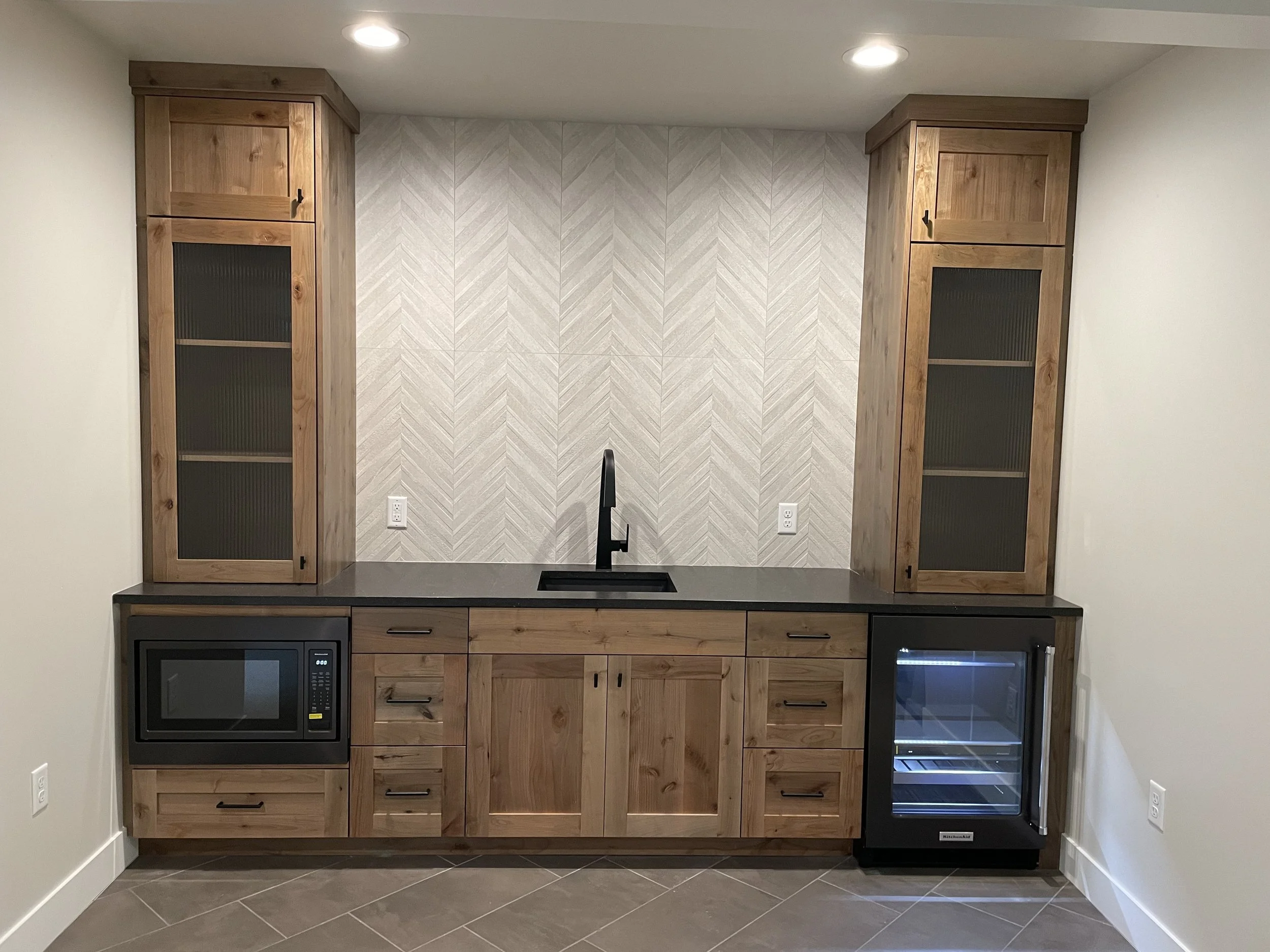 Kitchen area with wooden cabinets, a black microwave, a small wine fridge, and a textured tile backsplash.