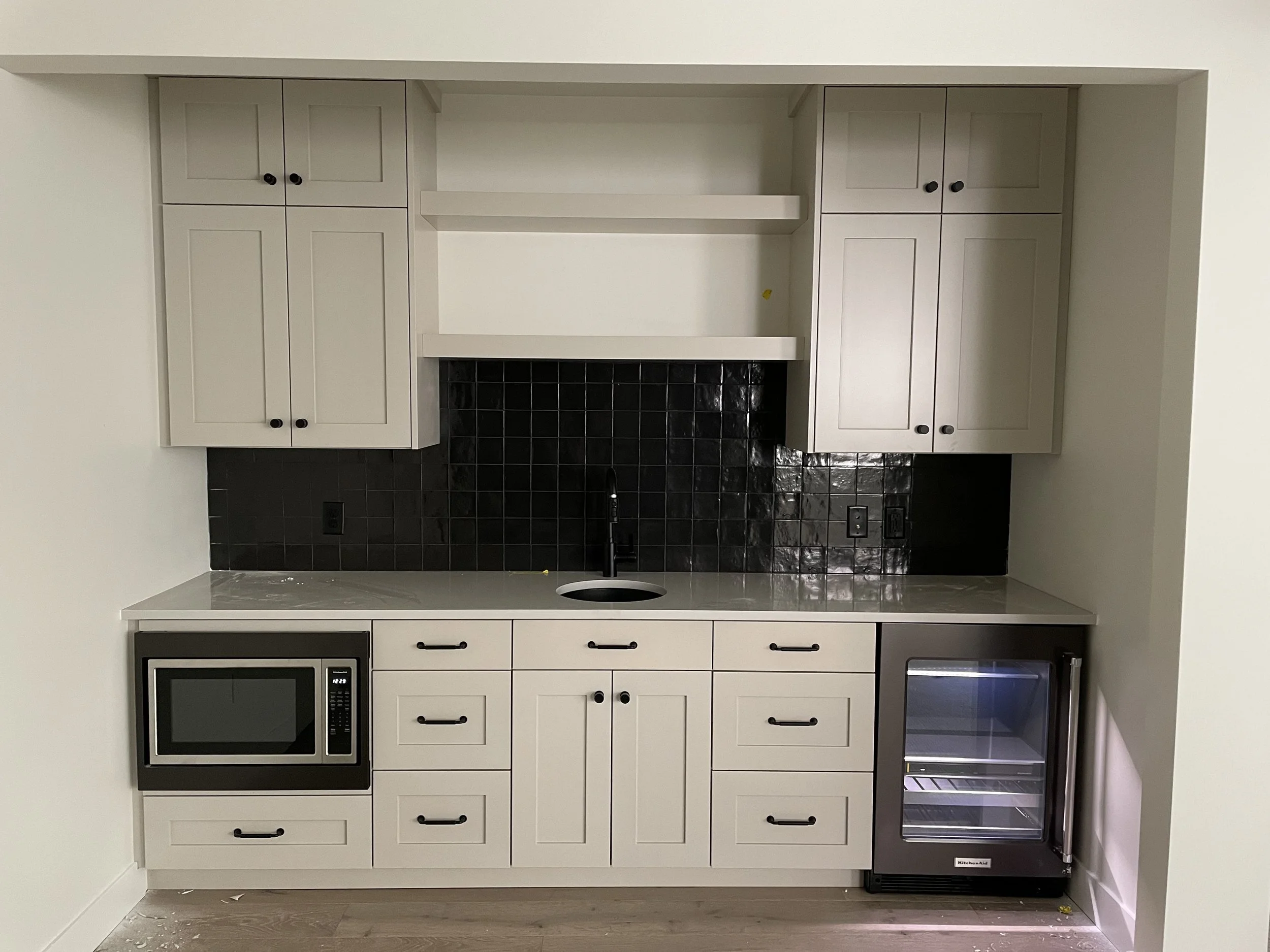 White kitchen cabinets with black handles surrounding a built-in space above a black tiled backsplash. There is a microwave oven in the lower left and a small stainless steel refrigerator in the lower right. The walls and ceiling are white, and the f