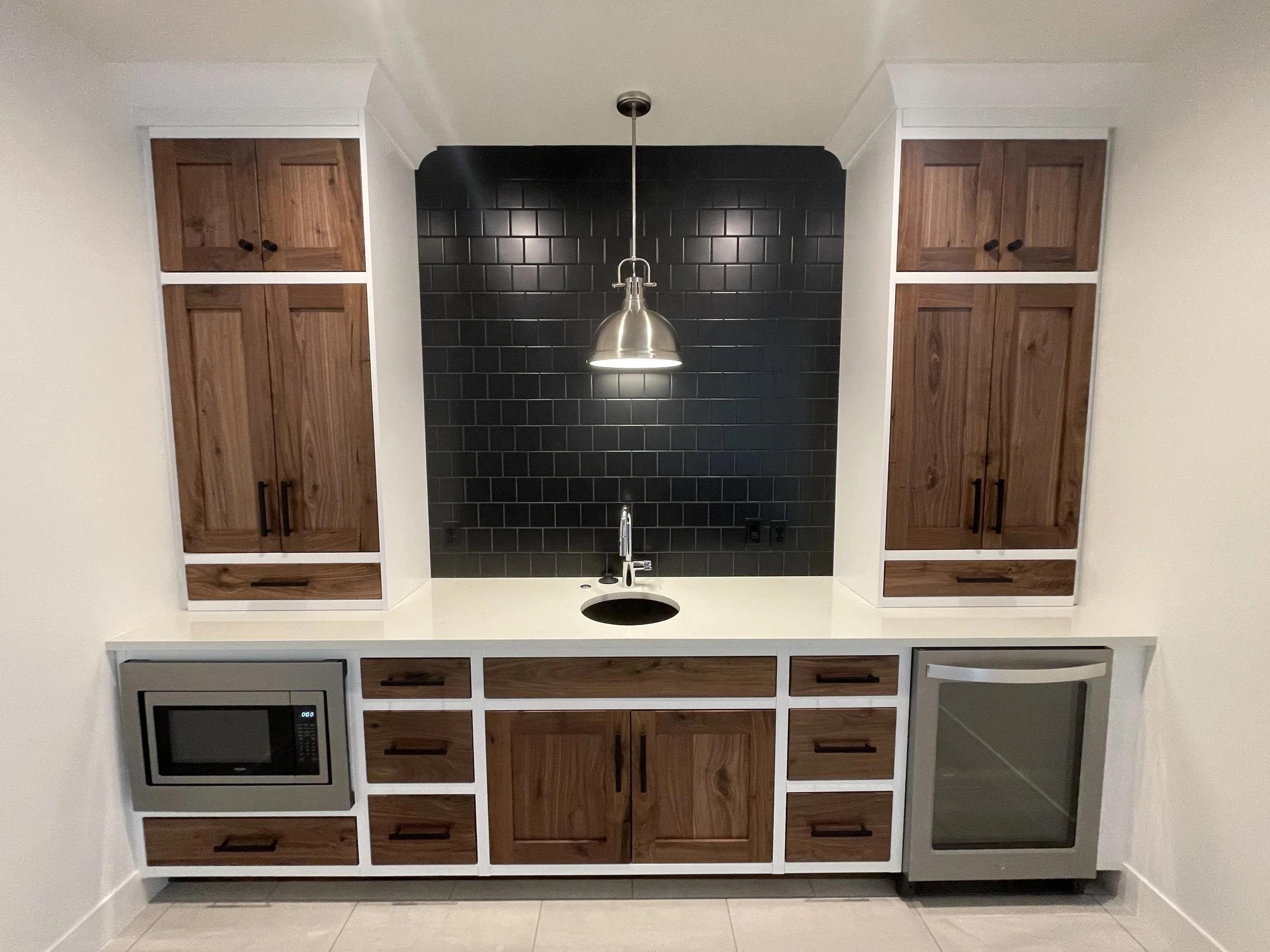 A kitchen with dark black tiled backsplash, white countertops, and wooden cabinets on both sides. There is a silver pendant light hanging from the ceiling over a sink, with various appliances and drawers below.
