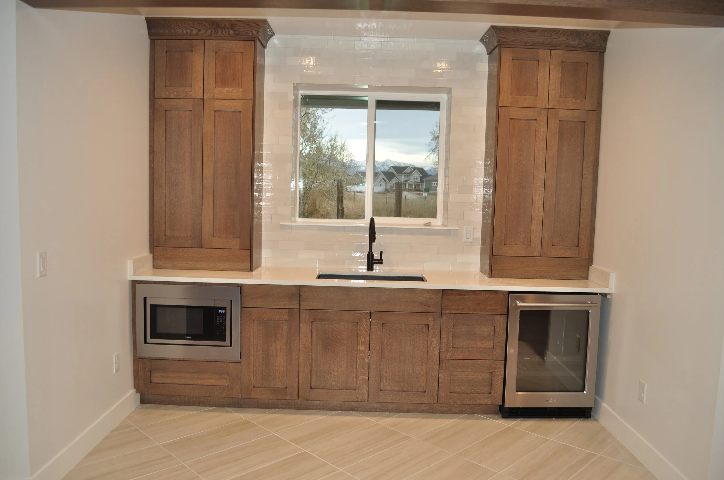 A modern kitchen with wooden cabinets, a black sink under a window, stainless steel oven and microwave, and tiled backsplash, overlooking a suburban neighborhood with mountains in the background.