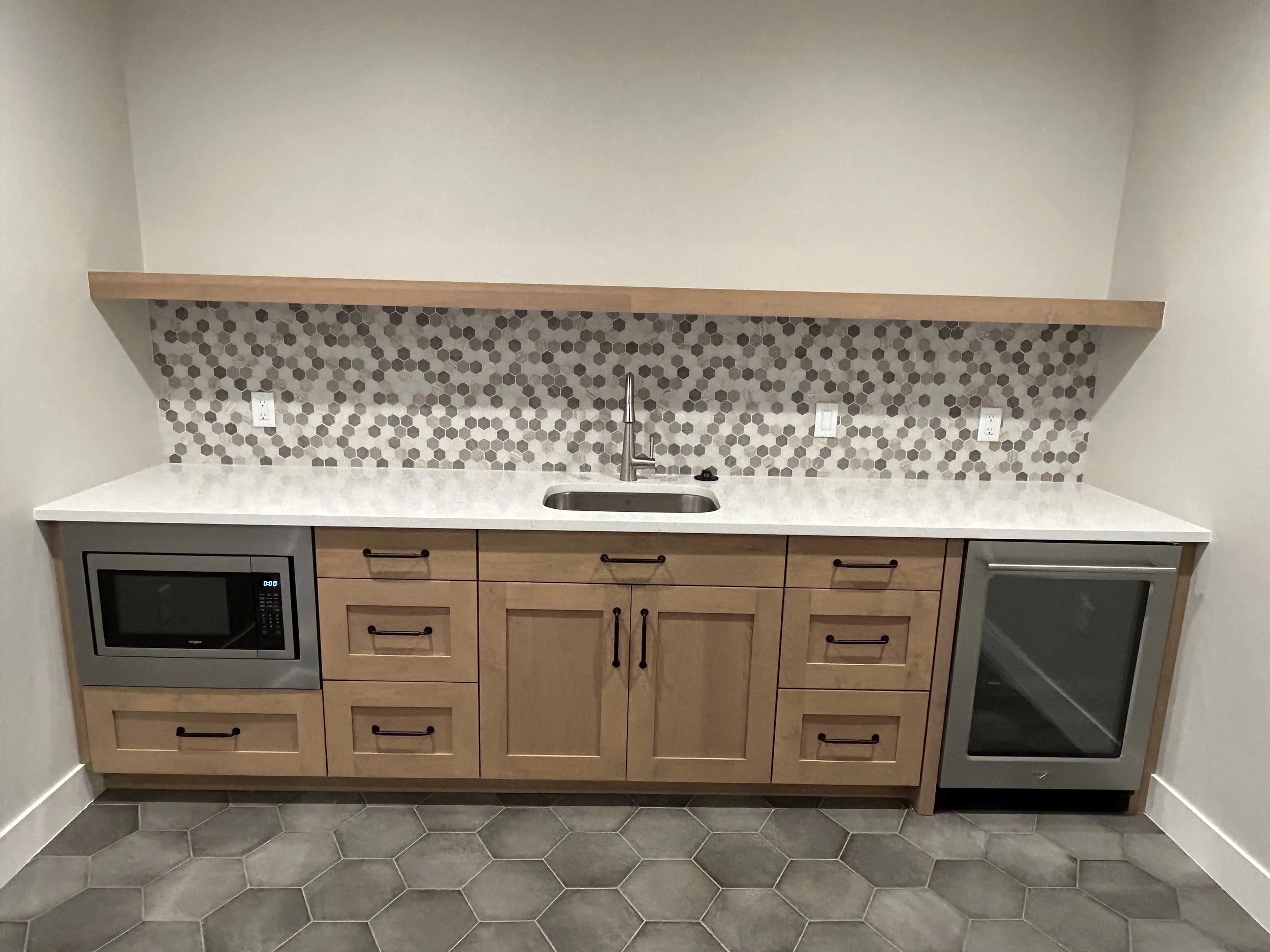 A kitchen countertop with a mosaic tile backsplash, a sink, and appliances including a microwave and oven cabinets above and below, with gray hexagonal floor tiles.