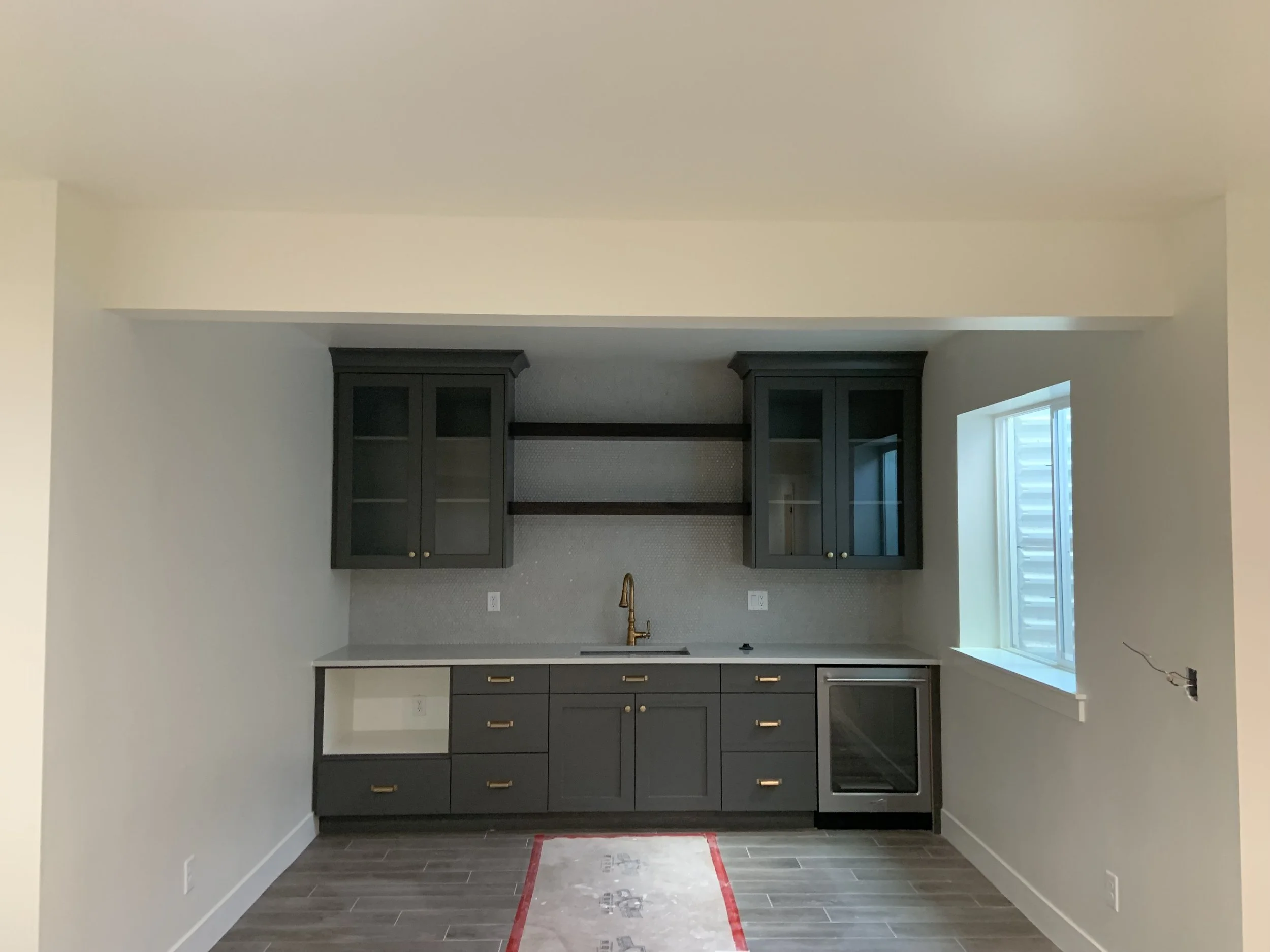 Modern kitchen with dark cabinetry, gray countertops, and brass handles, under construction renovation, with a window on the right and unfinished flooring.