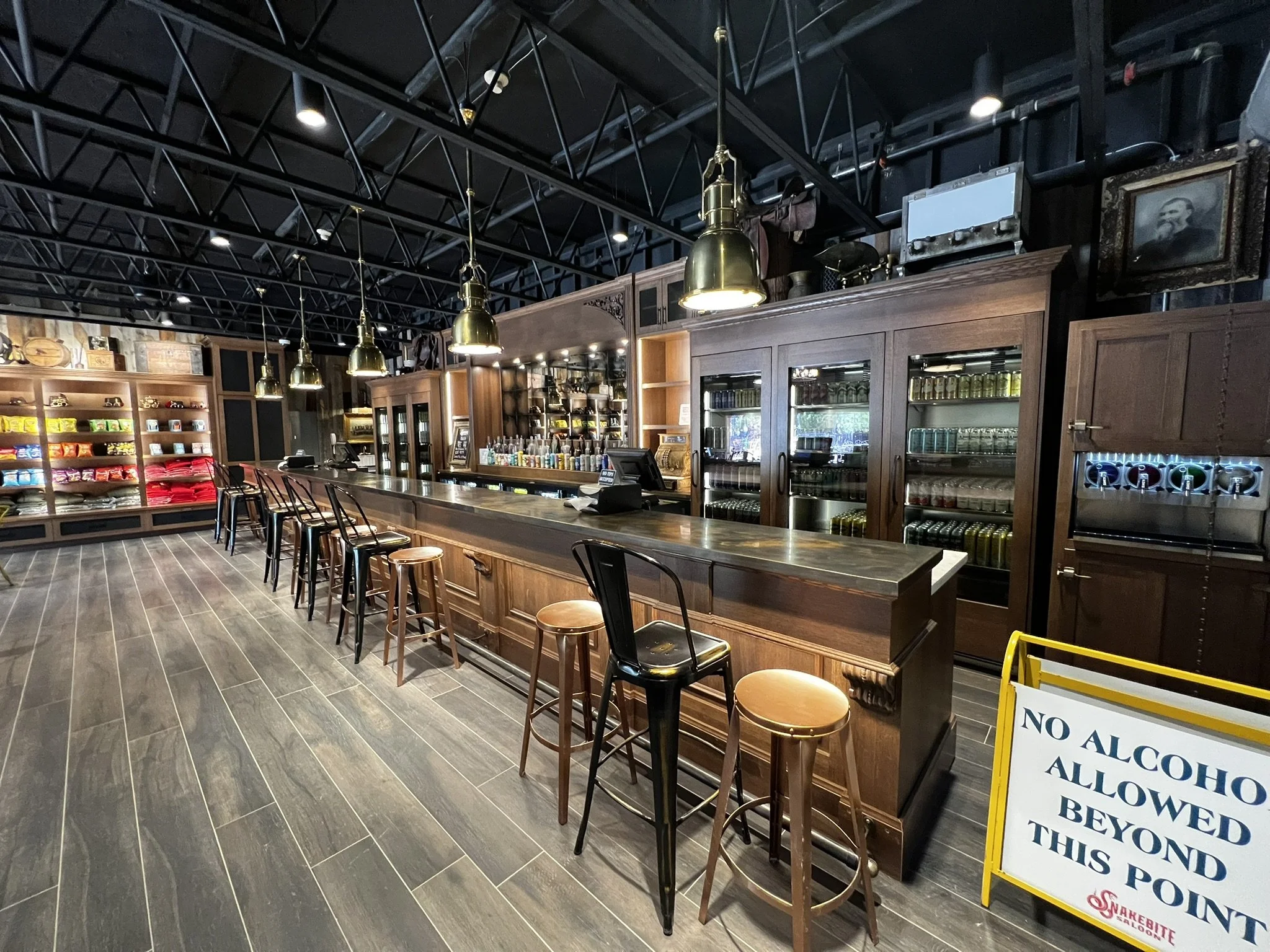 Interior of a bar or restaurant with dark wood furniture, a long bar counter, brass pendant lights, and shelves with colorful packaged products. There are chairs along the bar and a sign indicating a no alcohol policy beyond this point.