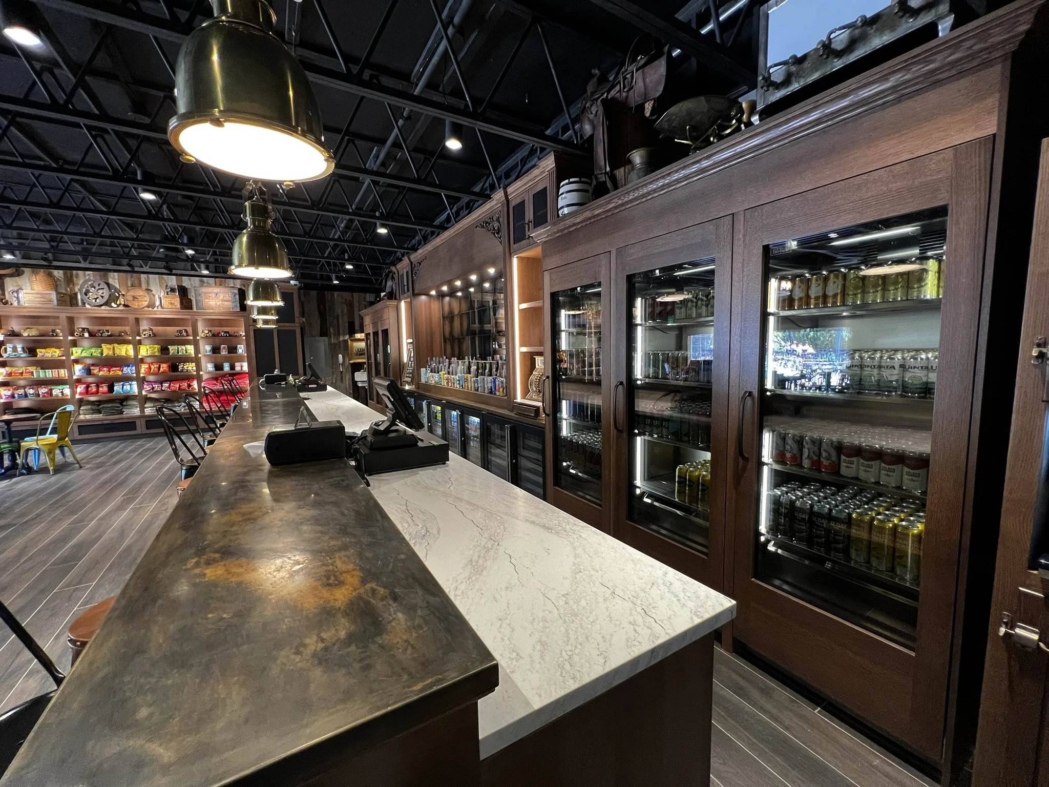 Counter at a coffee shop with a dark marble top, black chairs, and wooden shelves stocked with snacks and drinks, illuminated by large metallic pendant lights.
