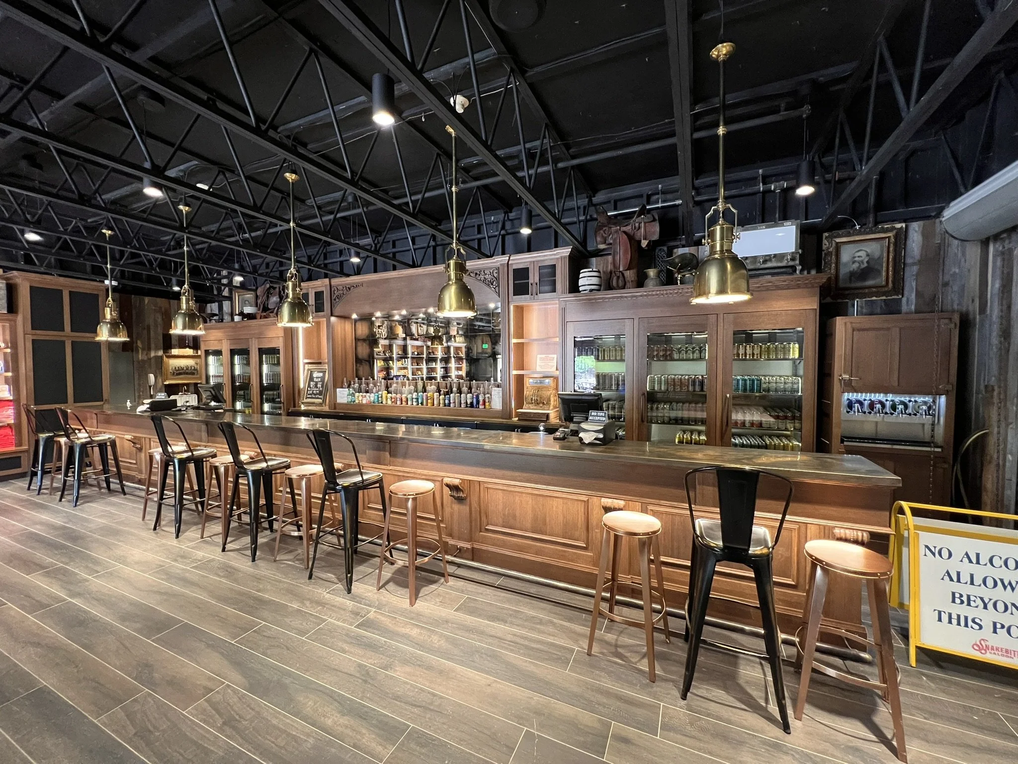 A modern bar interior with a long wooden bar counter, black barstools, and brass pendant lights hanging from a black ceiling with exposed trusses. There are wooden display cabinets behind the bar, stocked with bottles and cans, and framed pictures on