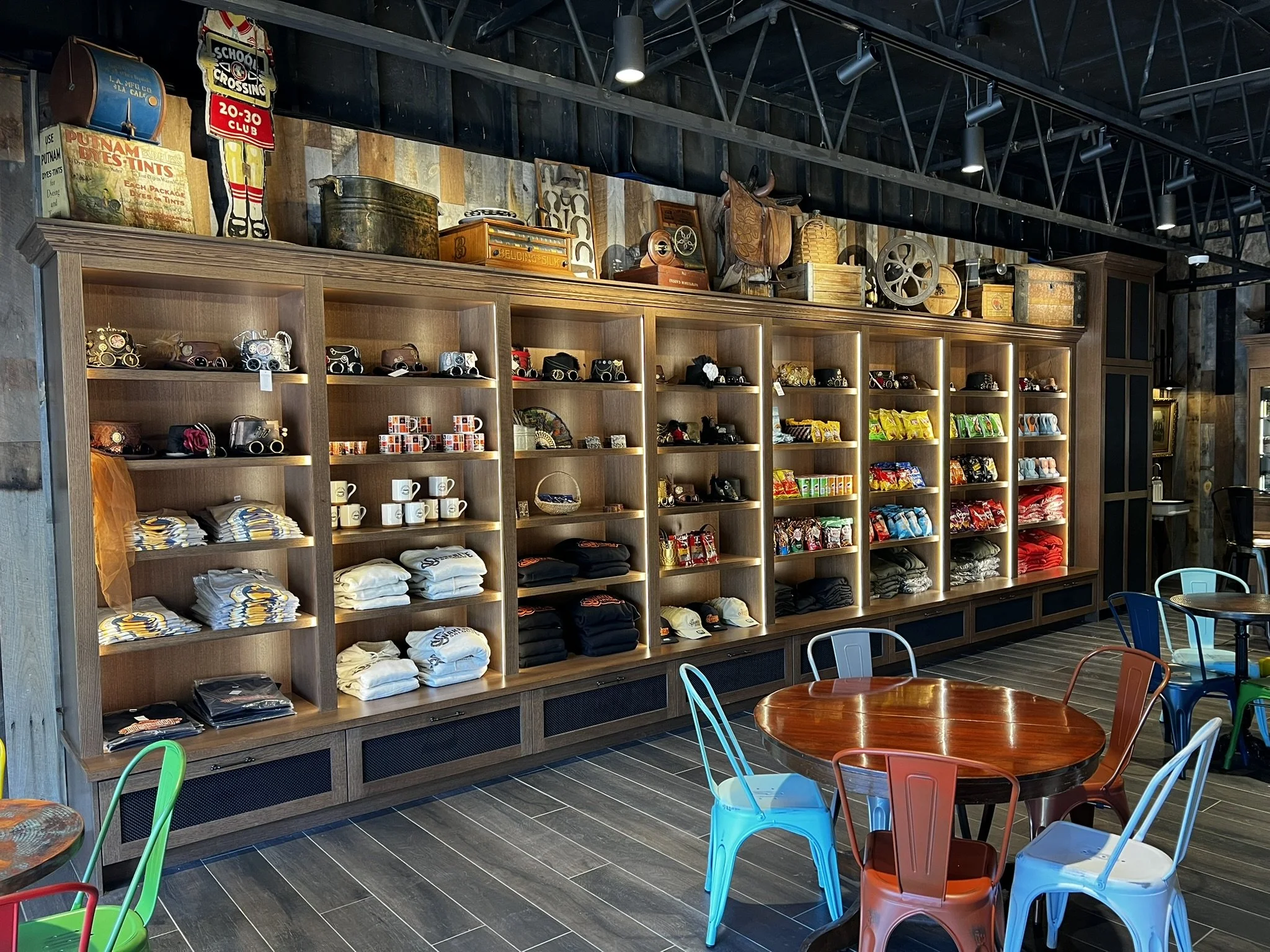 Interior of a retail store with wooden shelves displaying various merchandise including hats, mugs, snacks, and clothing. There are colorful chairs and tables in the foreground, and the back wall has vintage items and signs.