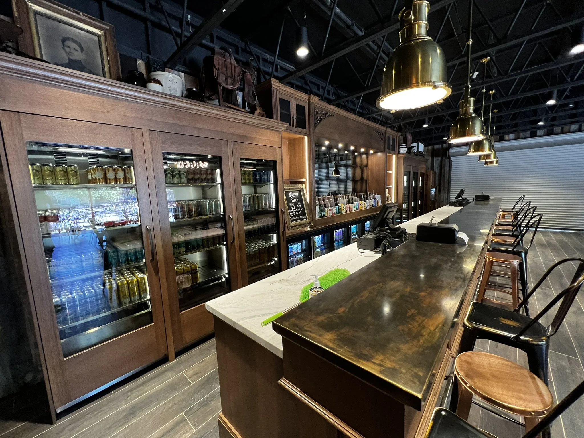 Inside a bar with made of dark wood, a row of golden hanging lamps, a bar counter with stools, a glass door fridge containing beverages, shelves with bottles and glasses, a chalkboard sign, and black ceiling with visible beams.