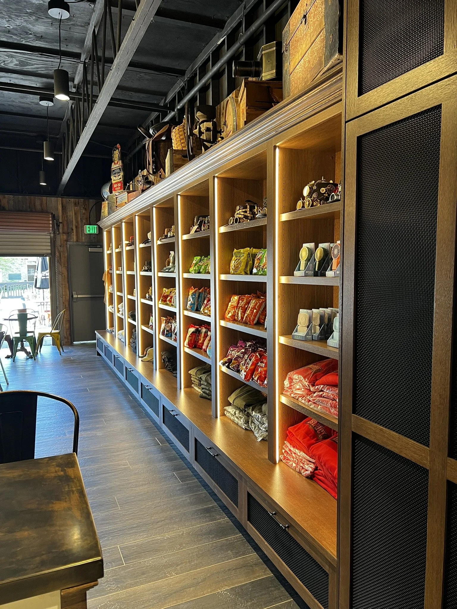 Interior of a retail store with wooden shelving filled with packaged snacks and small collectible toys. There are chairs near a large window with a view outside, and the ceiling has exposed beams and industrial lighting.