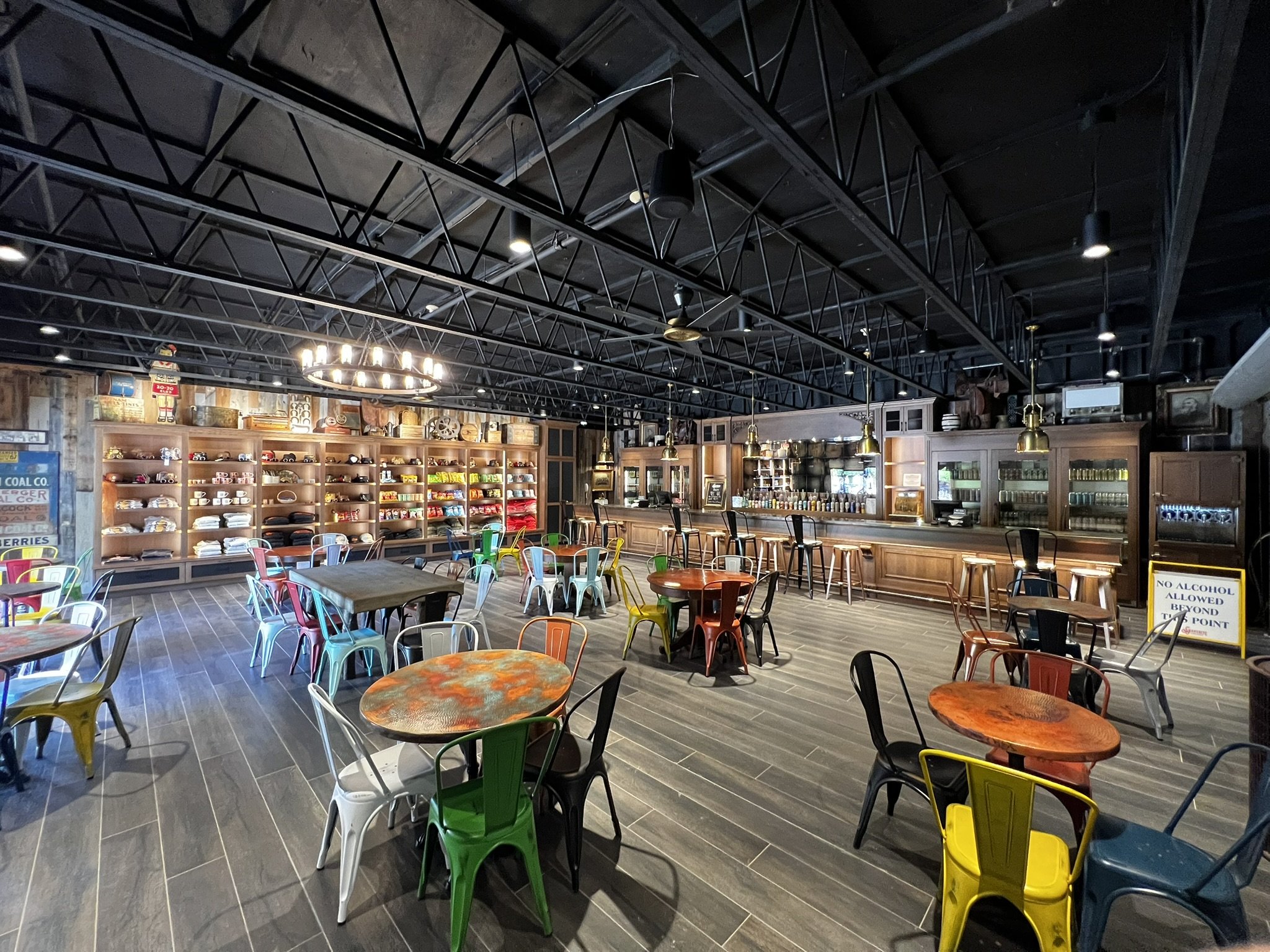 Inside a modern restaurant or cafe with colorful metal chairs, wooden tables, a wall with shelves filled with various mugs and decor, and a dark ceiling with exposed metal framing, lights, and ceiling fans.