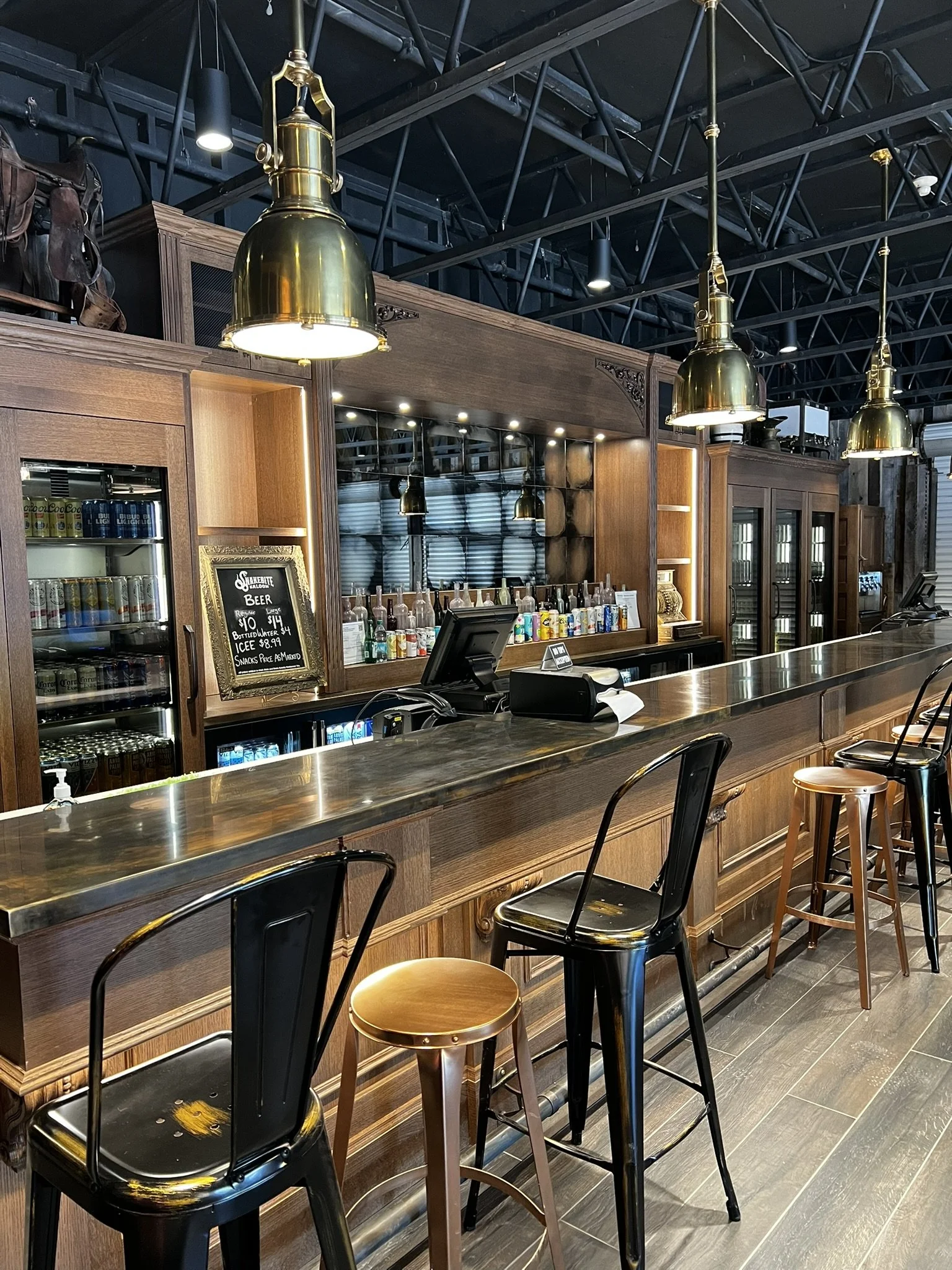 Inside a bar with wooden countertops and black chairs. Hanging brass pendant lights illuminate the area. Behind the bar, there are shelves with bottles and a chalkboard menu listing beer prices. The background features reflective glassware and a meta