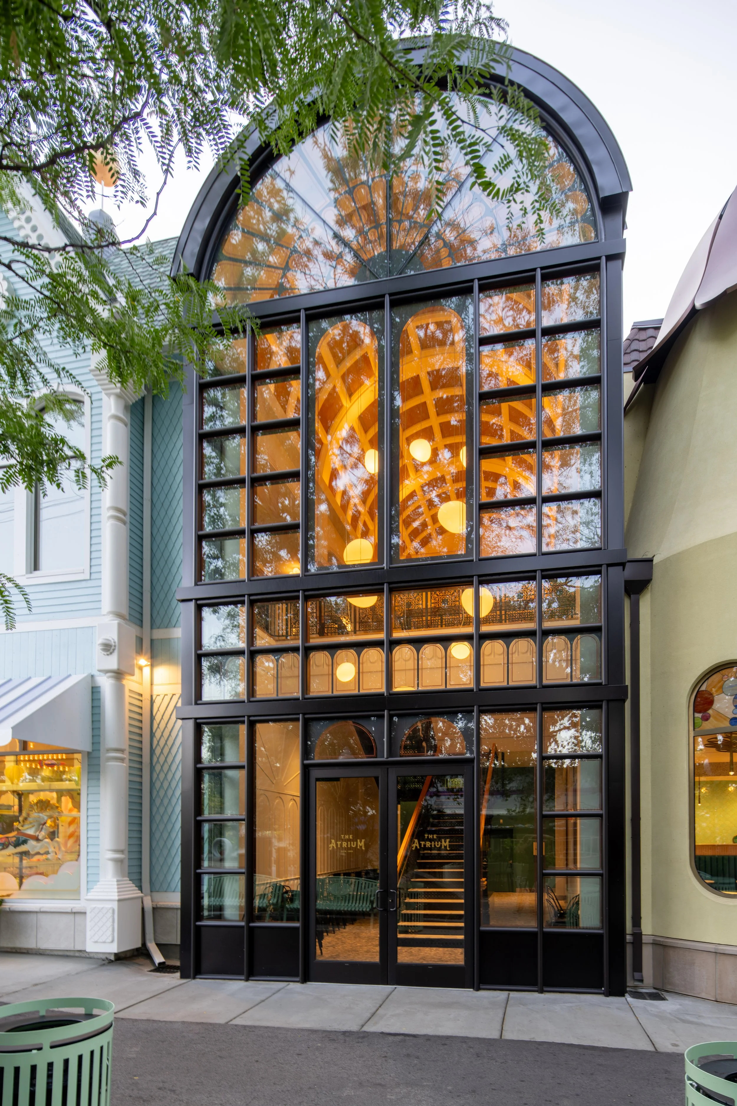 The image shows a contemporary building with a large glass window and arched roof, surrounded by greenery. The interior lights are on, illuminating the space inside.