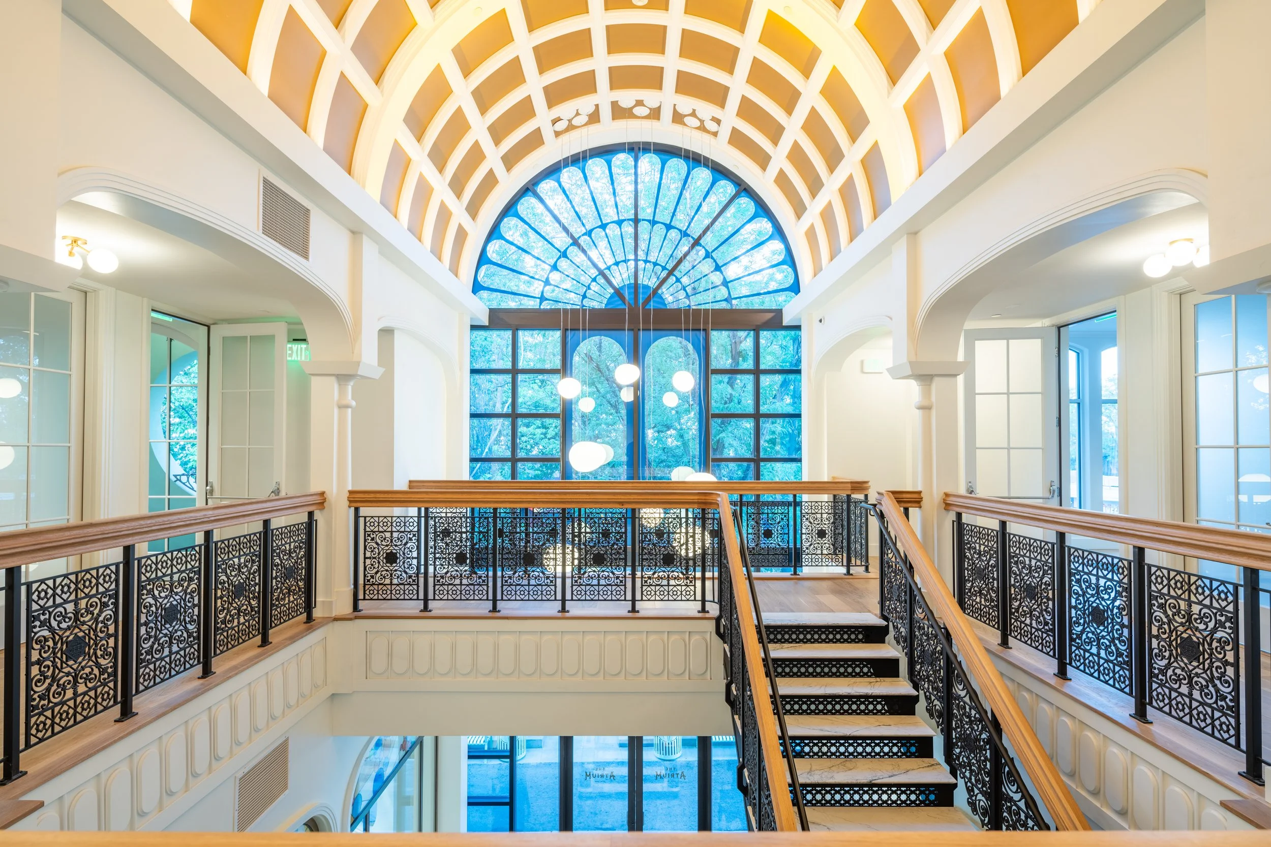 Interior view of a bright, modern building with large stained glass window archways, high vaulted ceiling with decorative grid pattern, elegant lighting fixtures, and a balcony with intricate wrought iron railing and wooden handrails.