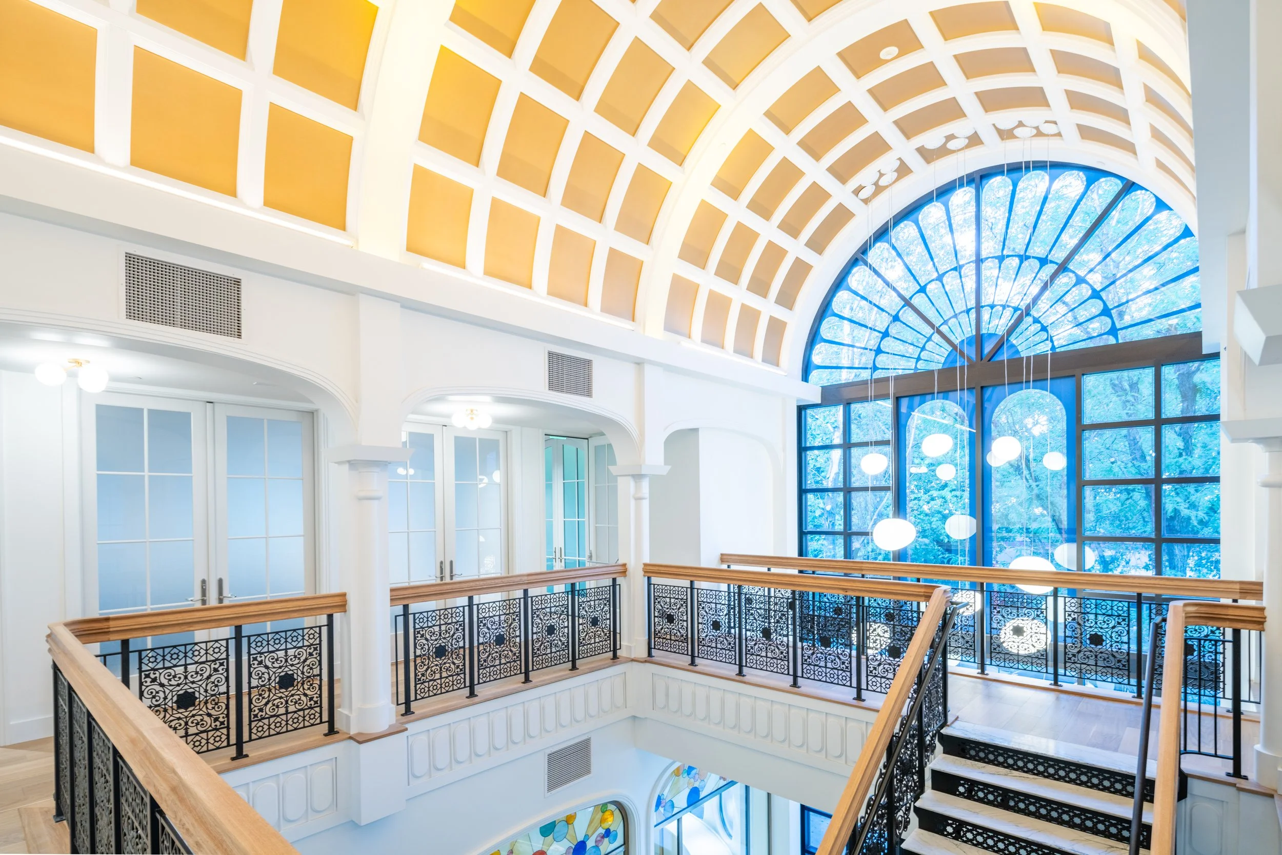Interior view of a building with a large stained glass window, a vaulted ceiling with yellow panels, and decorative railing along the upper level.