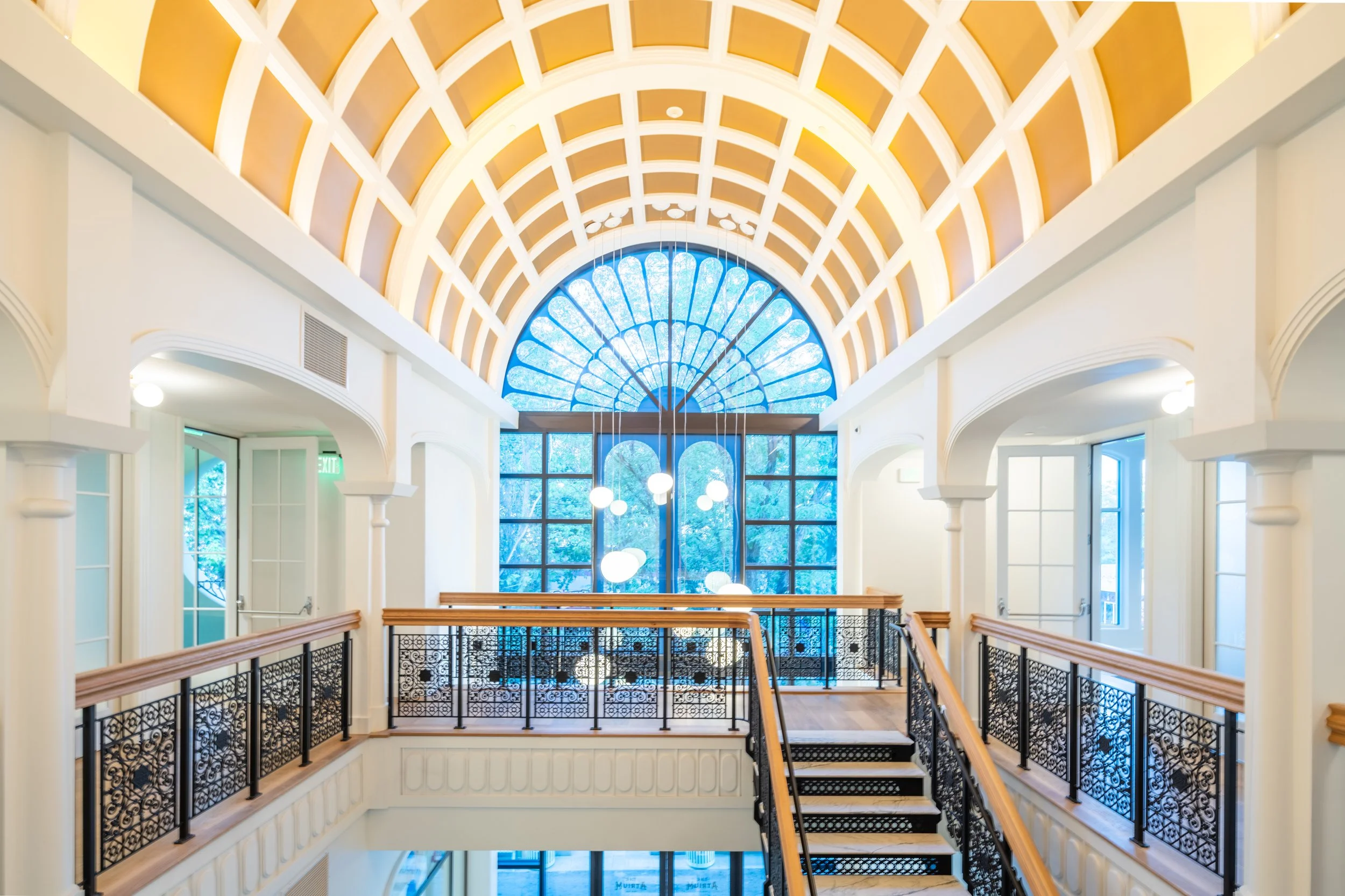 Interior of a building with a decorative arched ceiling featuring a large circular stained glass window at the end. The space has white walls, black wrought iron railings, and hanging circular light fixtures.