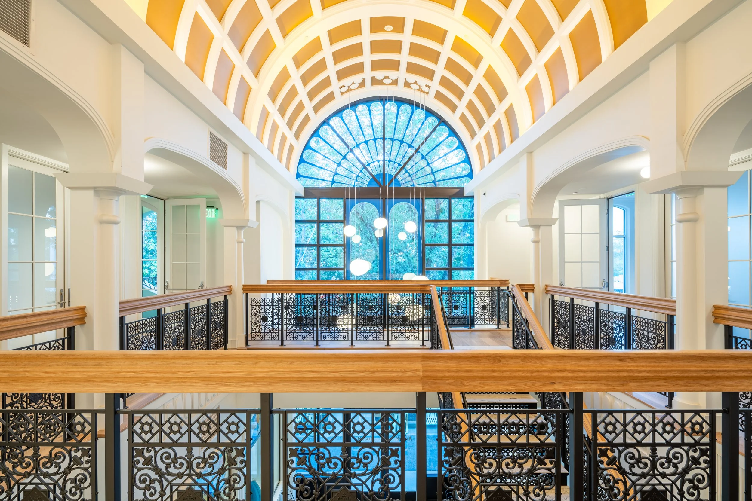Interior of a building with a large arched stained glass window, white walls, decorative black iron railings, wooden handrails, hanging lights, and a high vaulted ceiling with wooden beams.