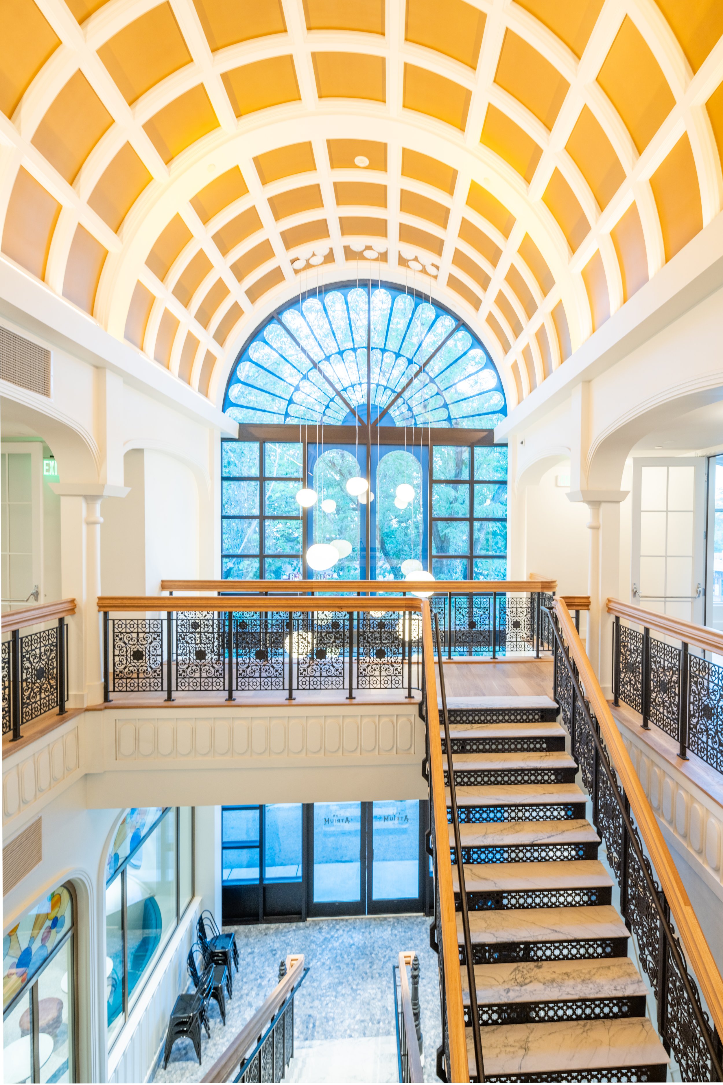 Interior of a building with a large arched window, a staircase with marble steps and ornate black railing, hanging lights, and decorative molding.