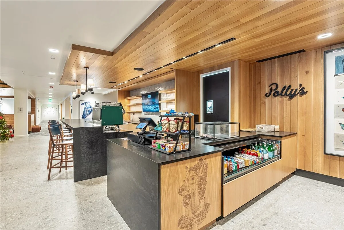 Interior view of a cafe or bar area with a wooden ceiling and wall. There is a black countertop with a cash register, snacks, and drinks. Shelves with glasses and a TV are visible behind the counter. The wall features the name 'Polly's' in black lettering.